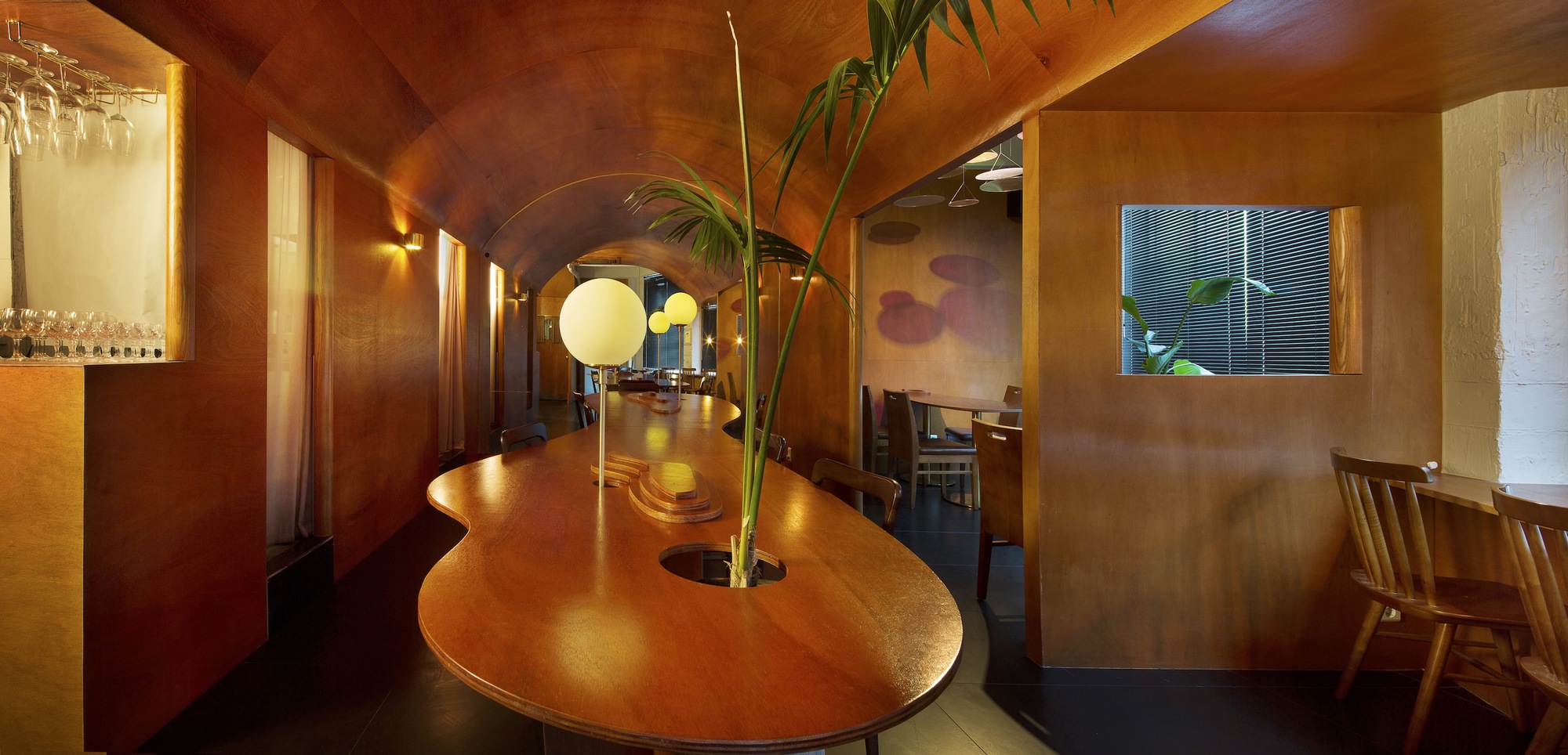 Vaulted timber ceiling over a curved dining table with palm fronds and globe pendant lights at dusk