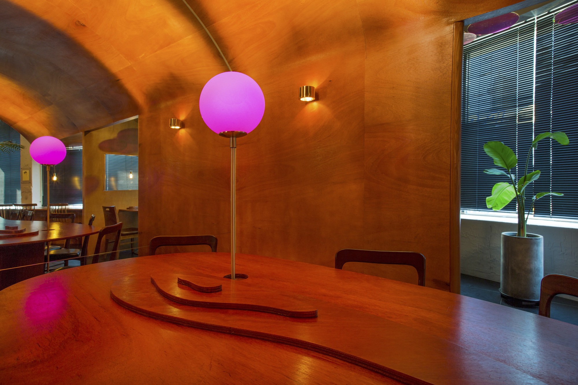 Dining area with curved timber table featuring inlaid grooves and illuminated pink globe fixtures under a vaulted ceiling