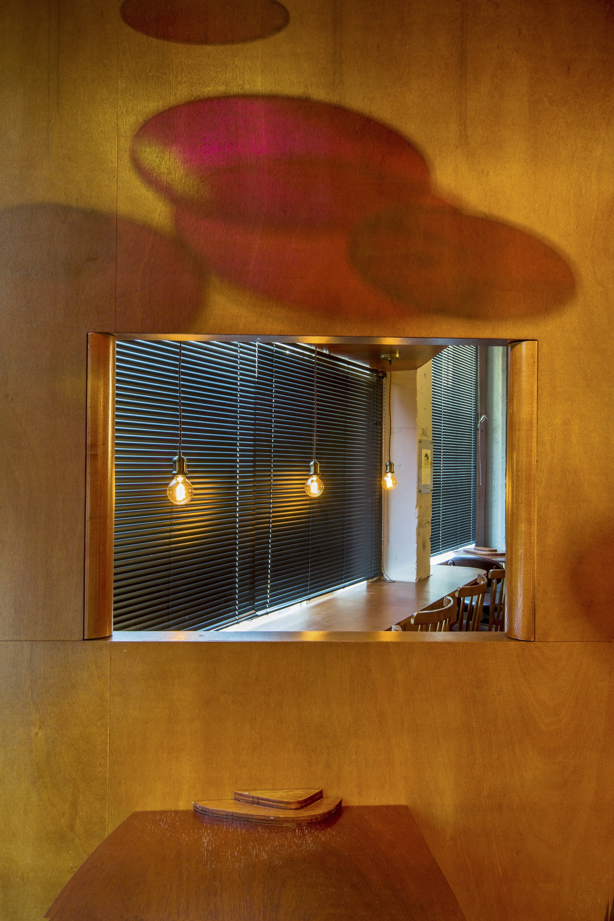 Framed aperture in wood-paneled wall revealing slatted metal screens and pendant lights with reflected colored light shadows