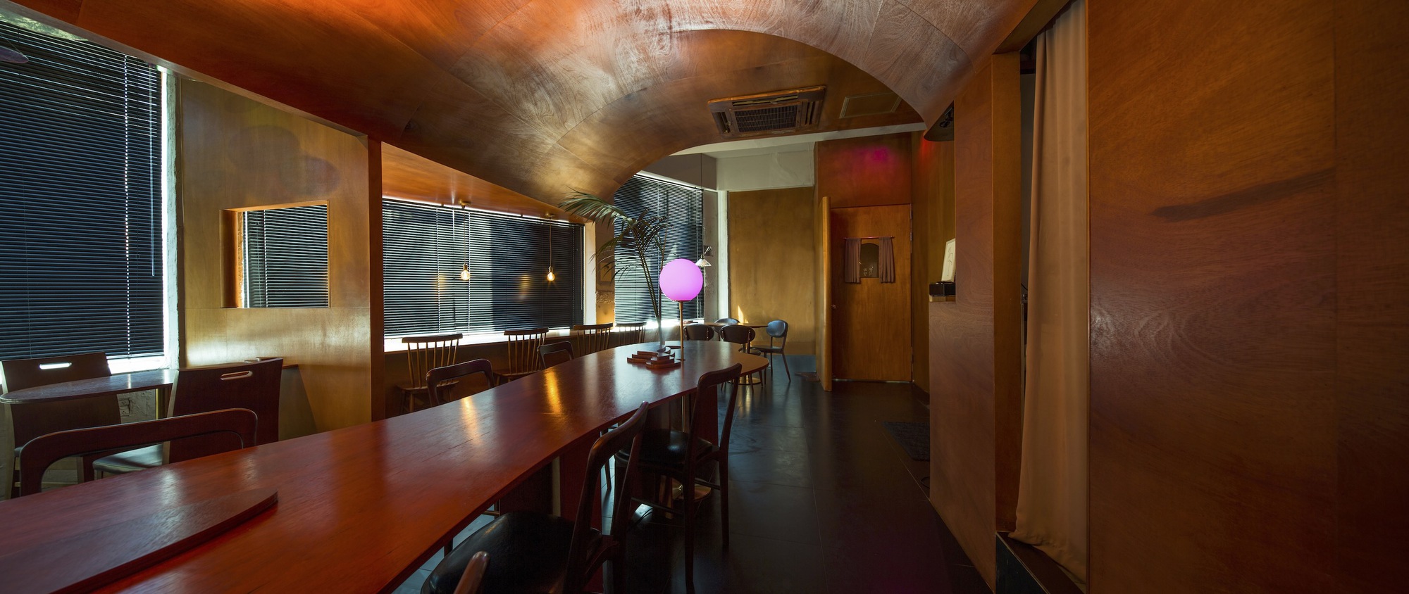 Panoramic dining hall with barrel-vaulted timber ceiling, horizontal blinds, and long wooden tables lit by pink globe lamp