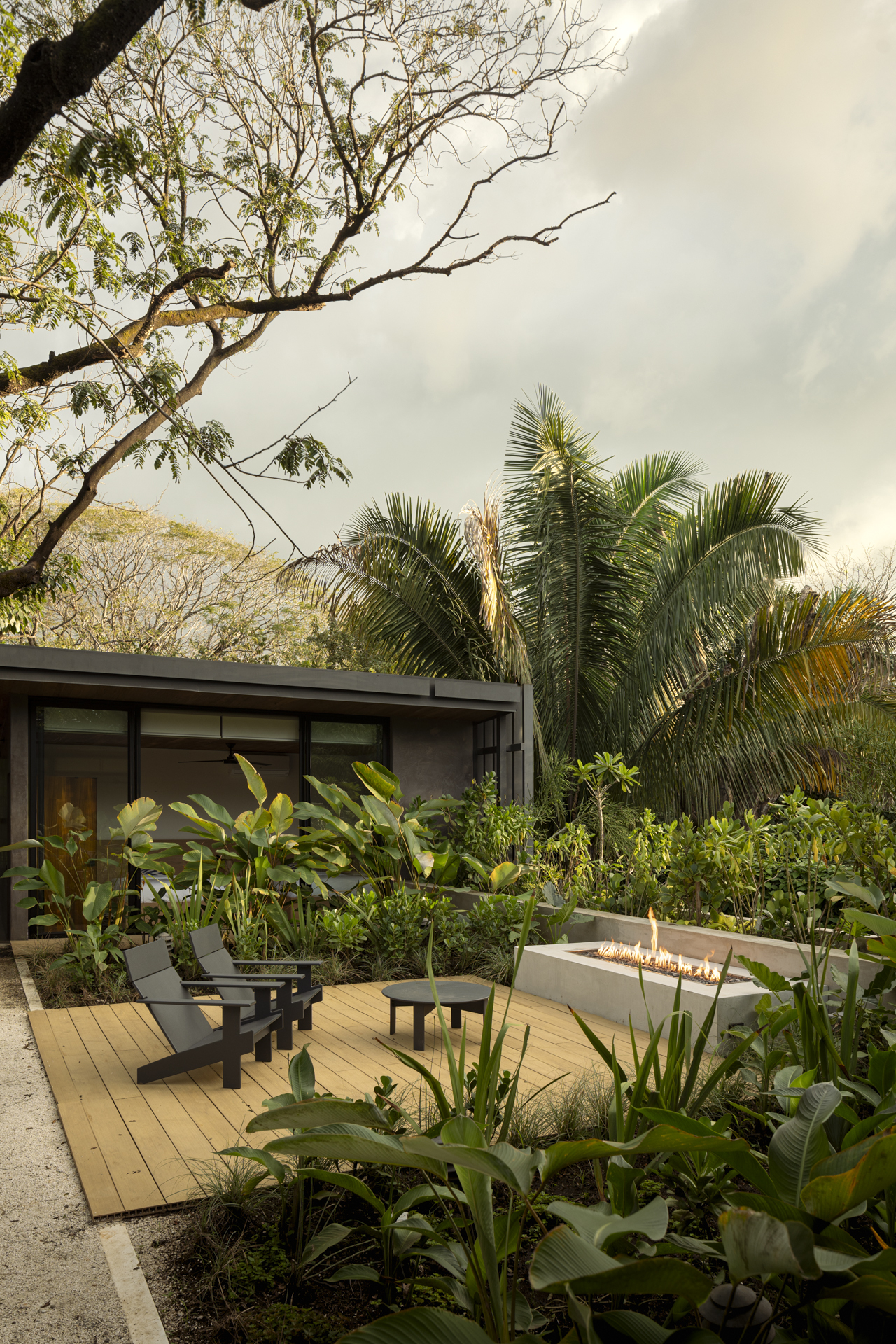 Timber deck terrace with fire feature and lounge chairs amid planted beds under overcast sky