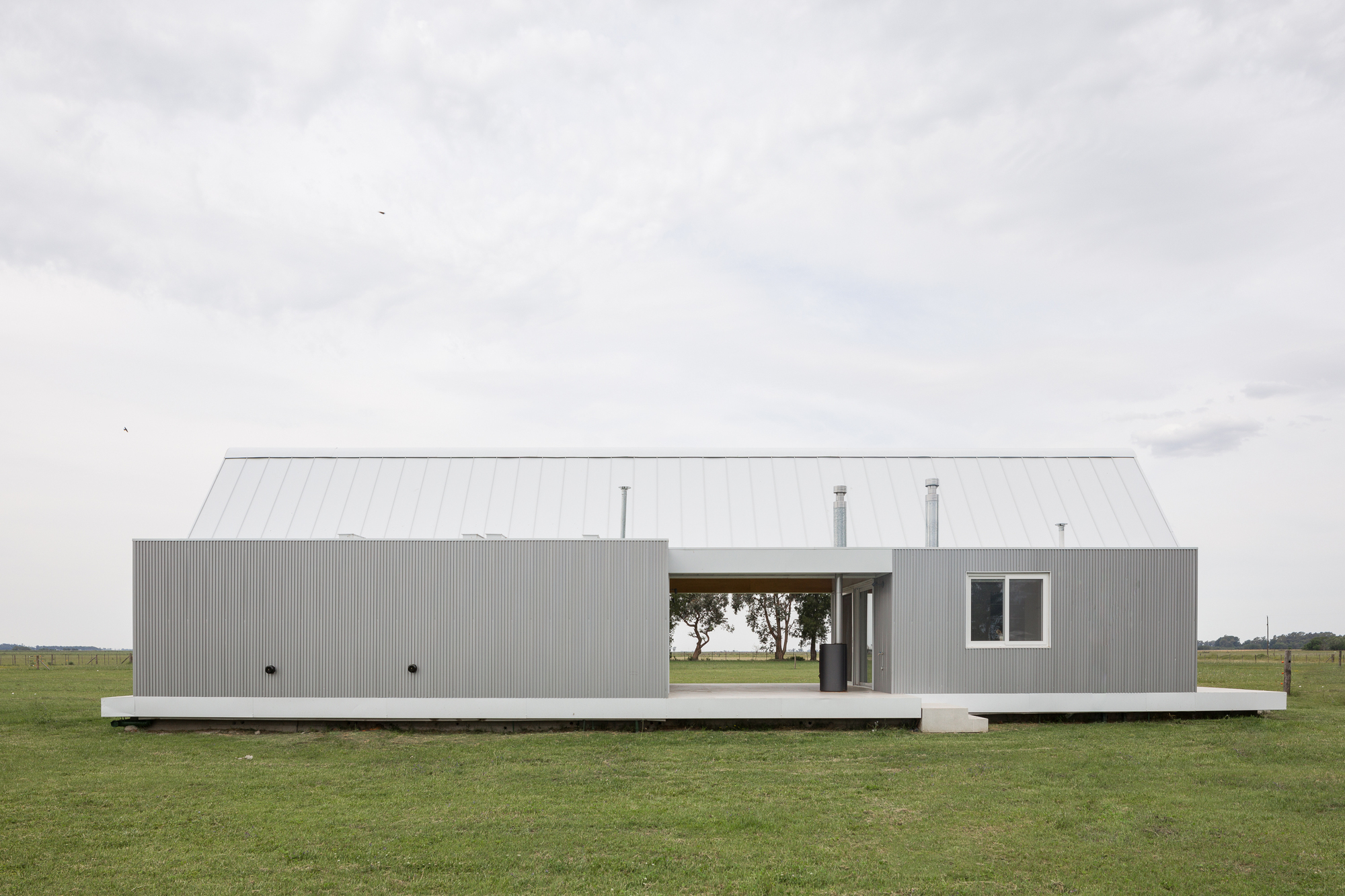 Long side elevation of corrugated metal volume on concrete plinth in grassy field