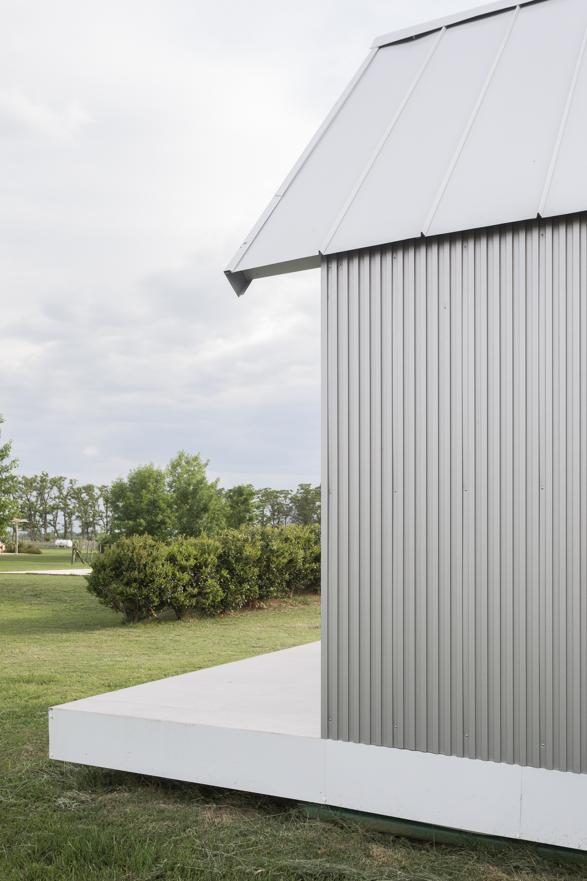 Corrugated metal volume with white plinth base and angled roof plane against a cloudy sky