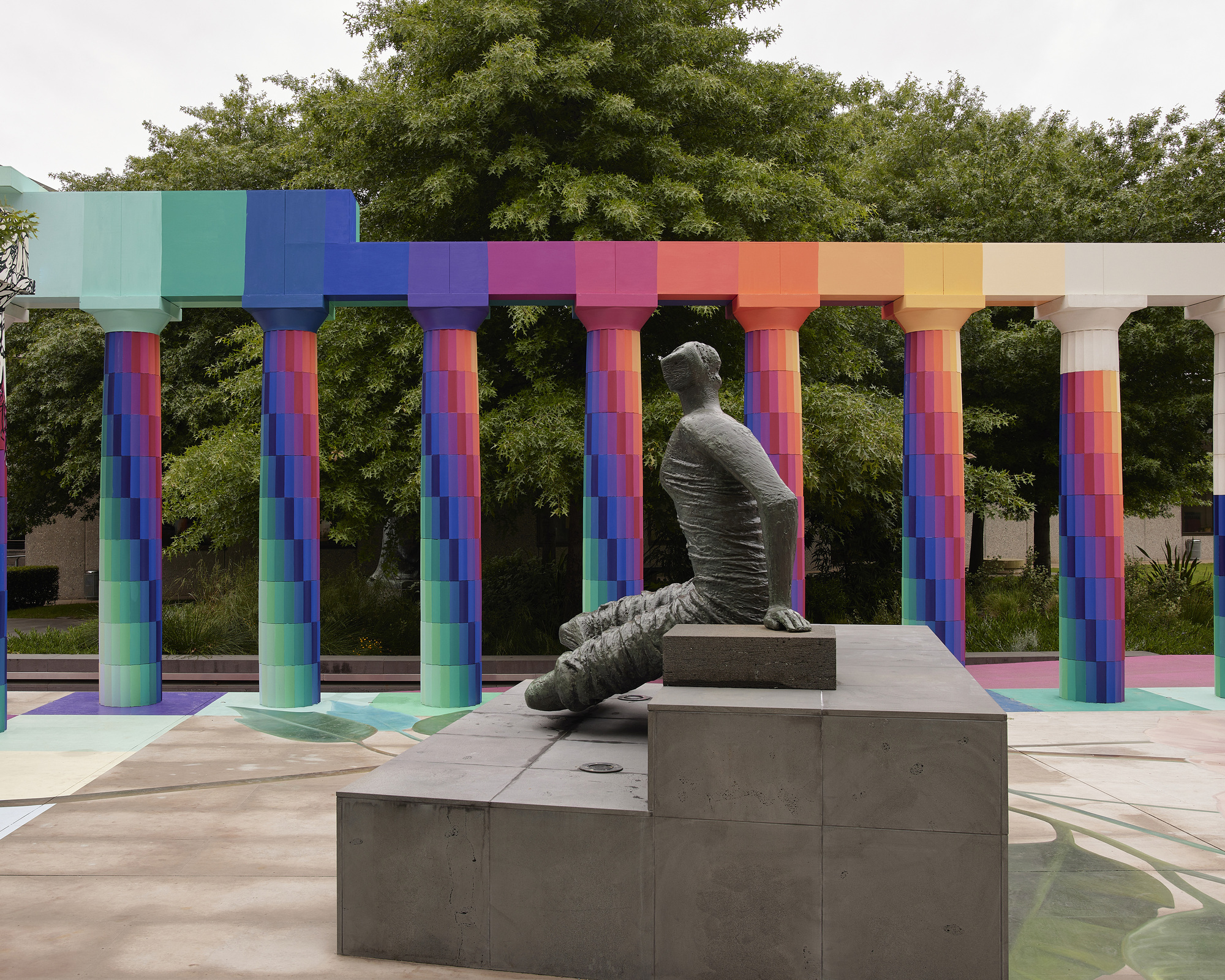 Concrete sculpture of a seated figure on stepped plinth beneath the rainbow-gradient column capitals and beam