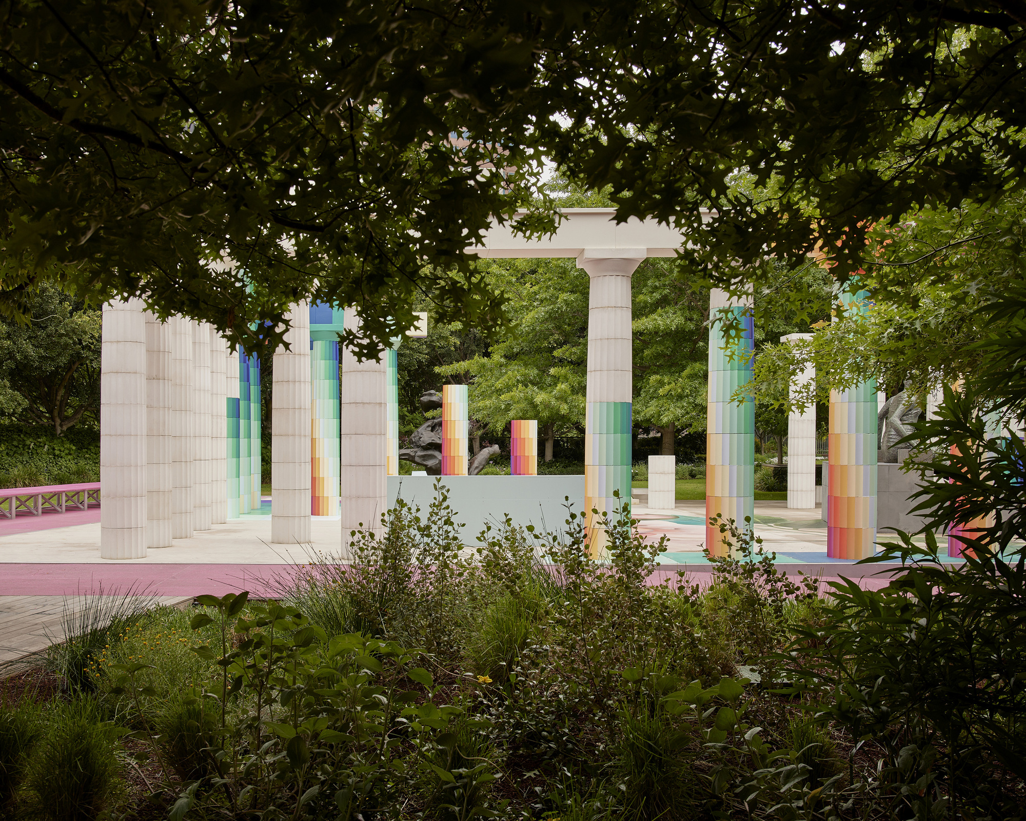 Open-air pavilion framed by mature tree canopy with pixelated columns and pink paving visible beyond