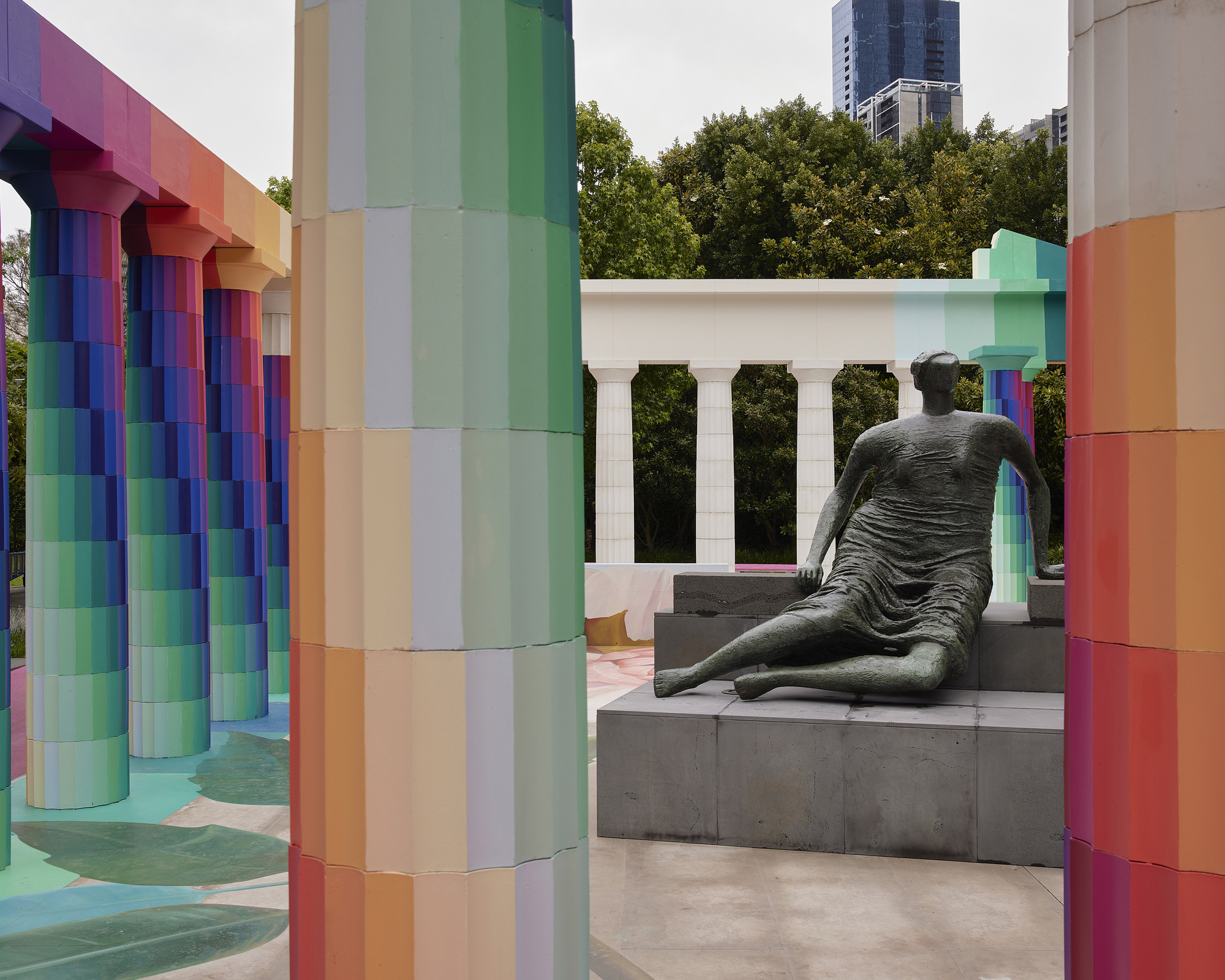 Rainbow gradient tiled columns framing a bronze sculpture on grey stone steps with white colonnade beyond