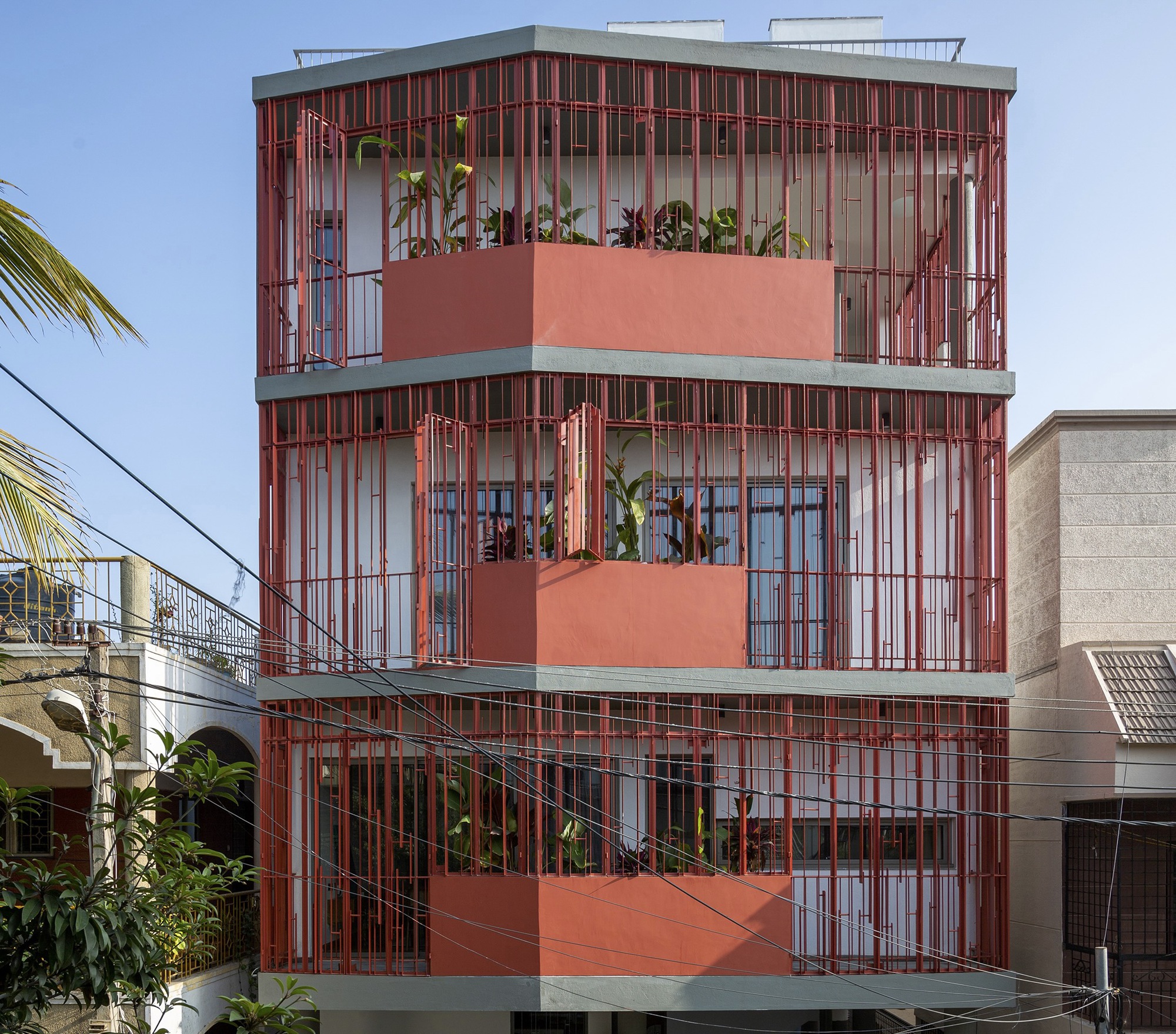 Street facade with red vertical metal brise-soleil wrapping three levels and planted terrace balconies