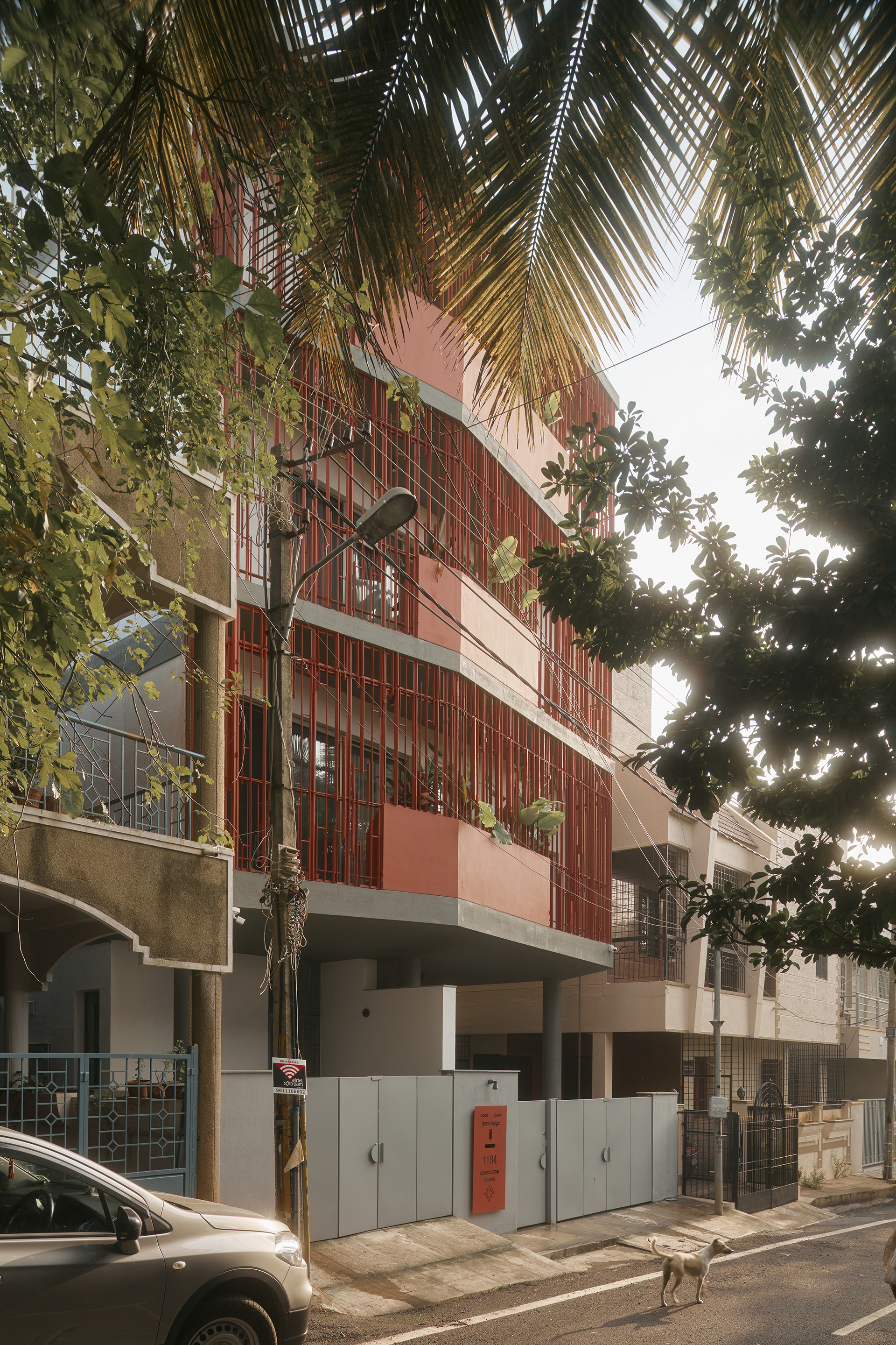 Street view of the red vertical batten facade rising above palm fronds and parked vehicles