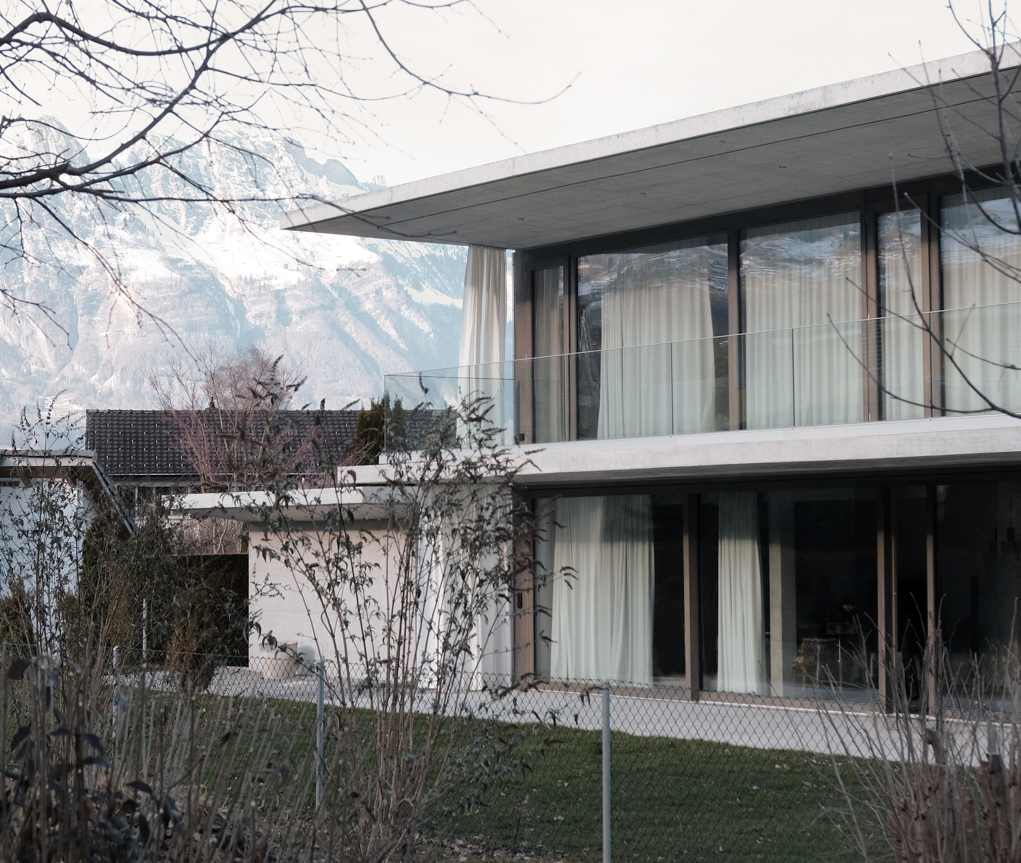 Two-storey concrete and glass facade with deep overhangs framed by bare winter branches and distant mountains