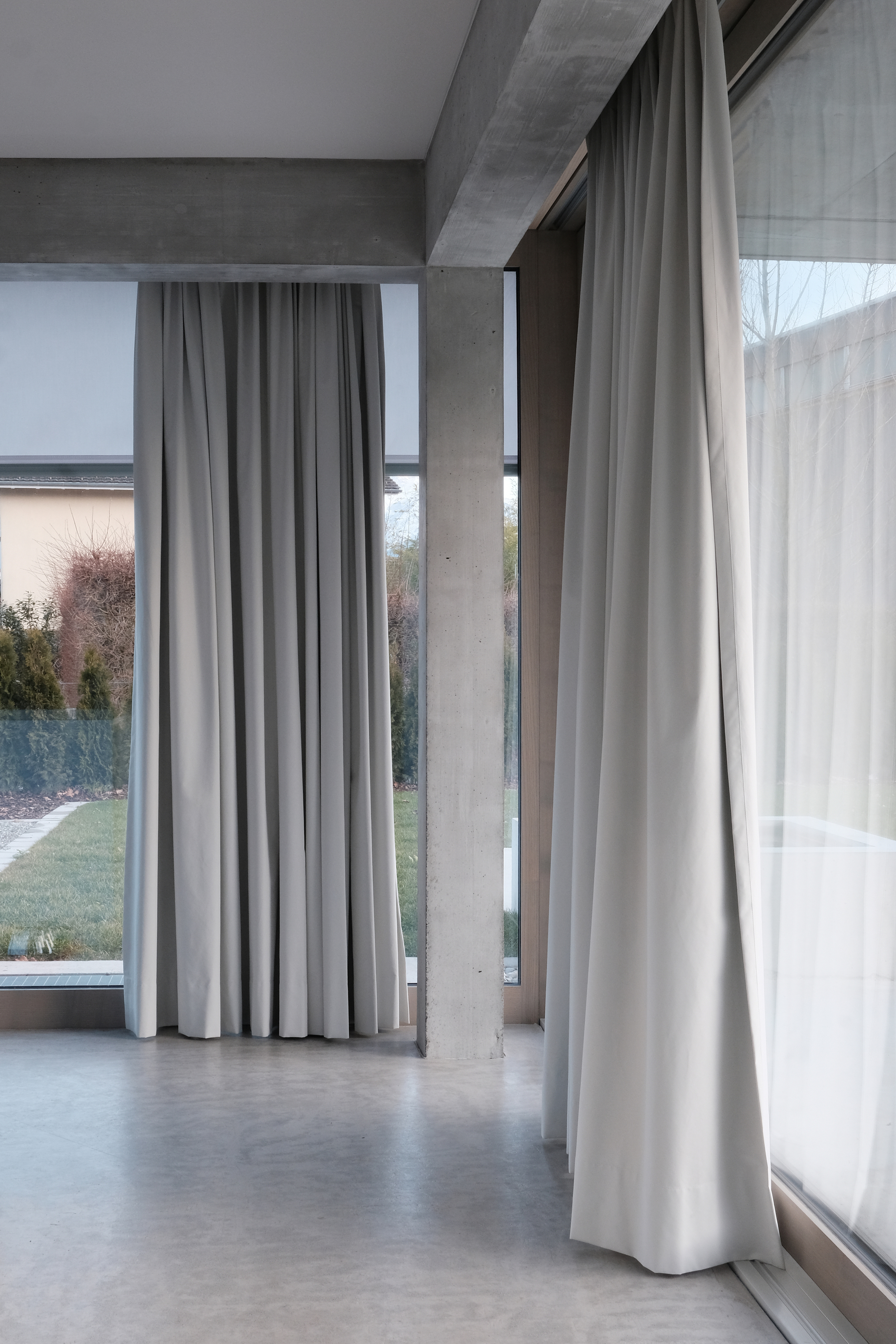 Interior view of full-height curtains along concrete framed windows opening to a garden in twilight