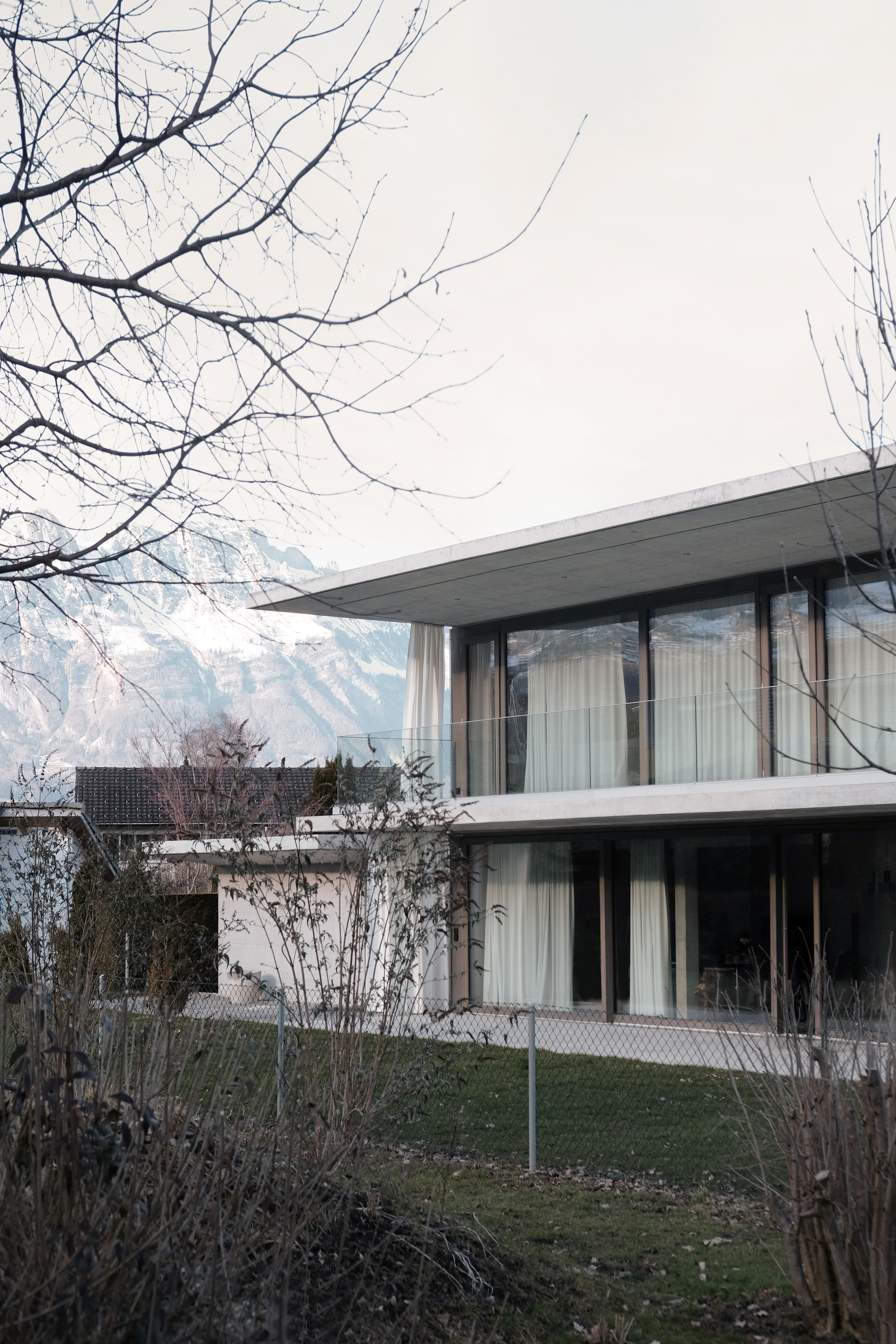 Two-story concrete volume with floor-to-ceiling glazing and sheer curtains framed by bare winter branches