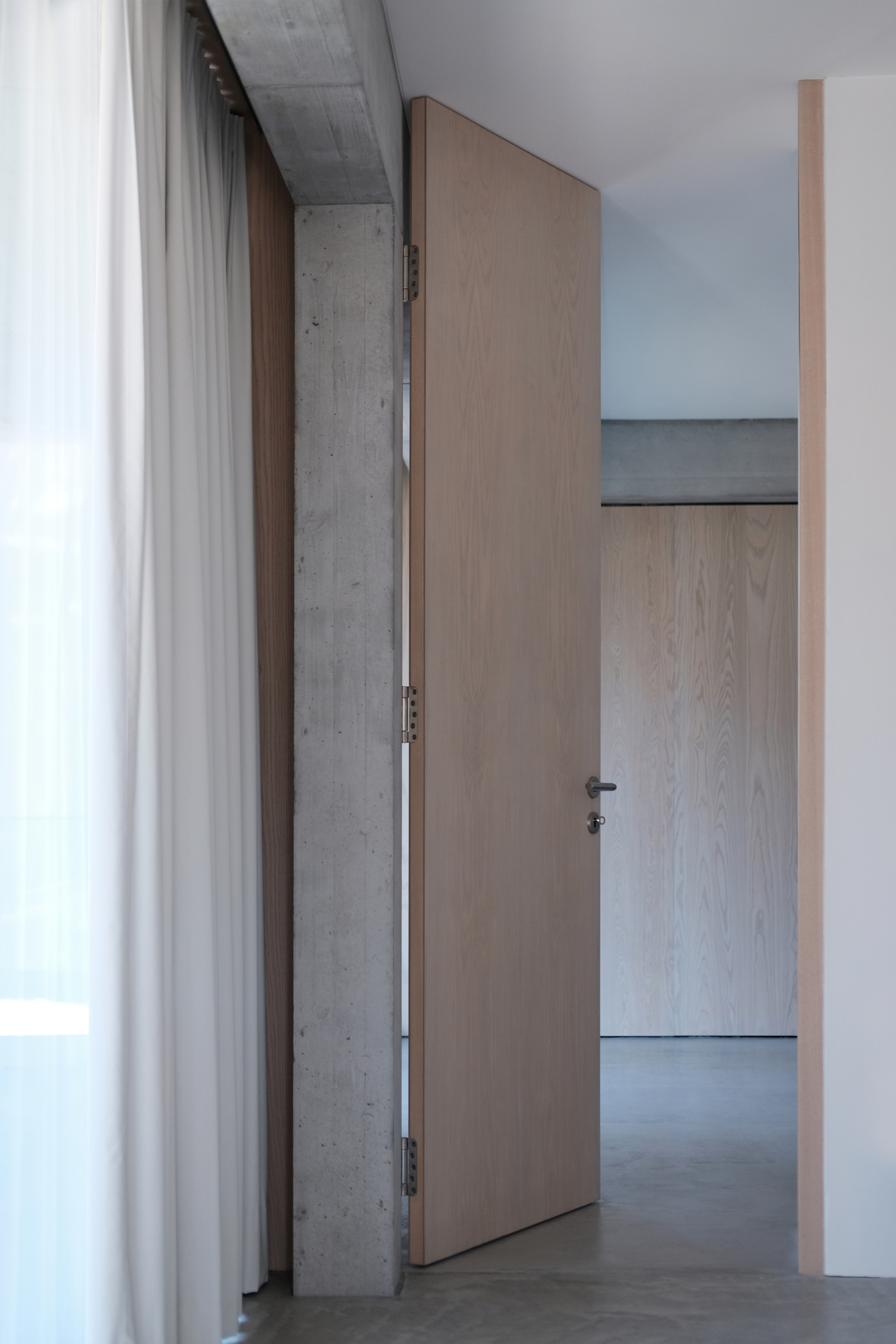 Interior hallway with light wood door ajar against concrete beam and white curtain at left