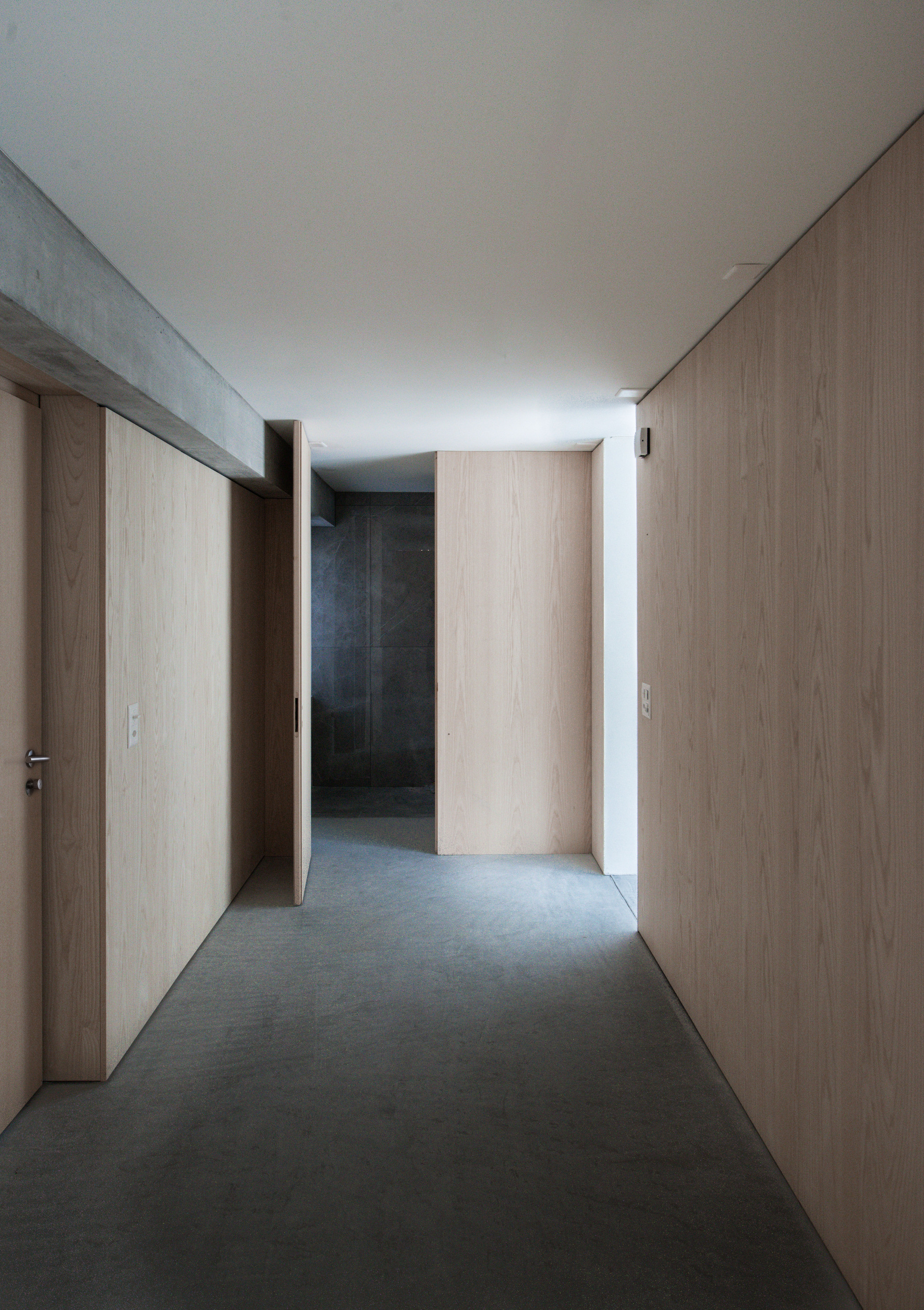 Narrow corridor with pale wood walls and concrete beams leading to backlit darker passage ahead