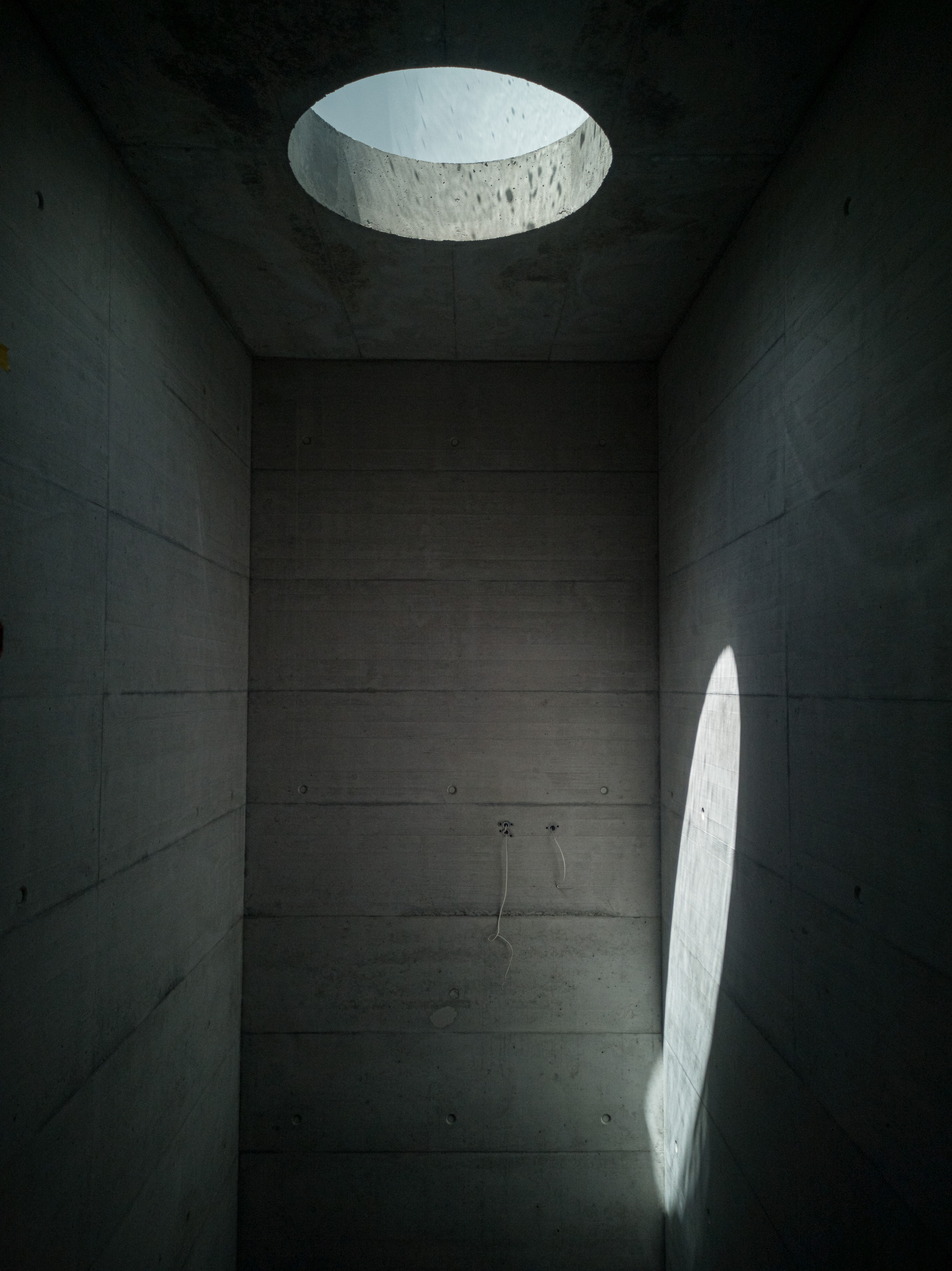 Board-formed concrete bathing chamber lit by circular skylight and arched wall opening