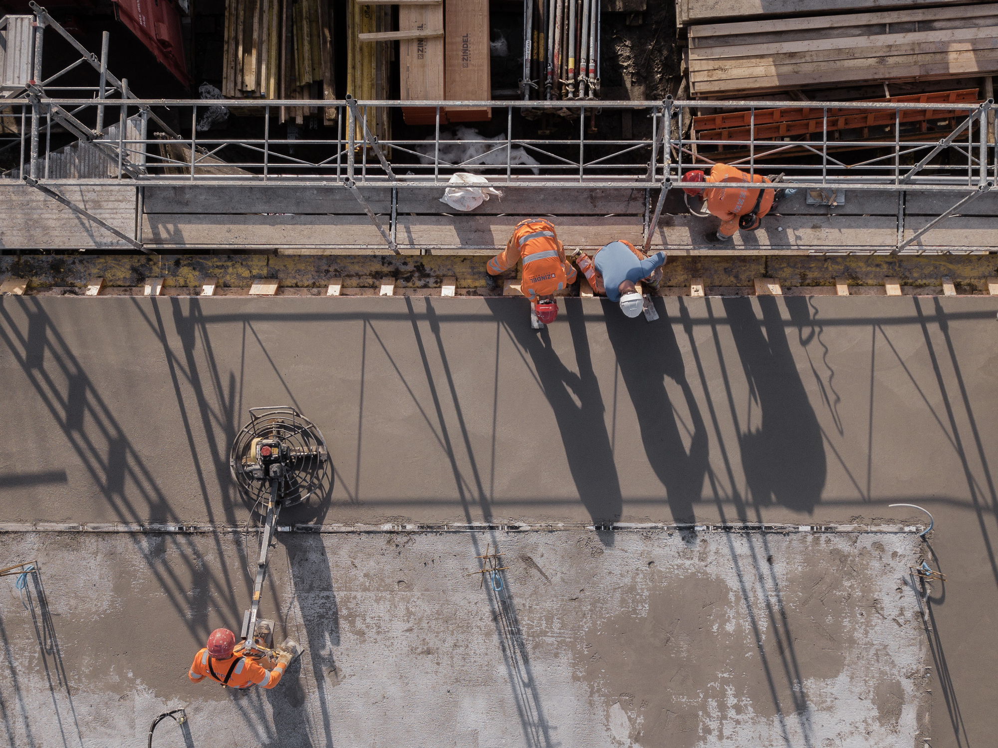 Construction workers on fresh concrete slab casting shadows through scaffolding above
