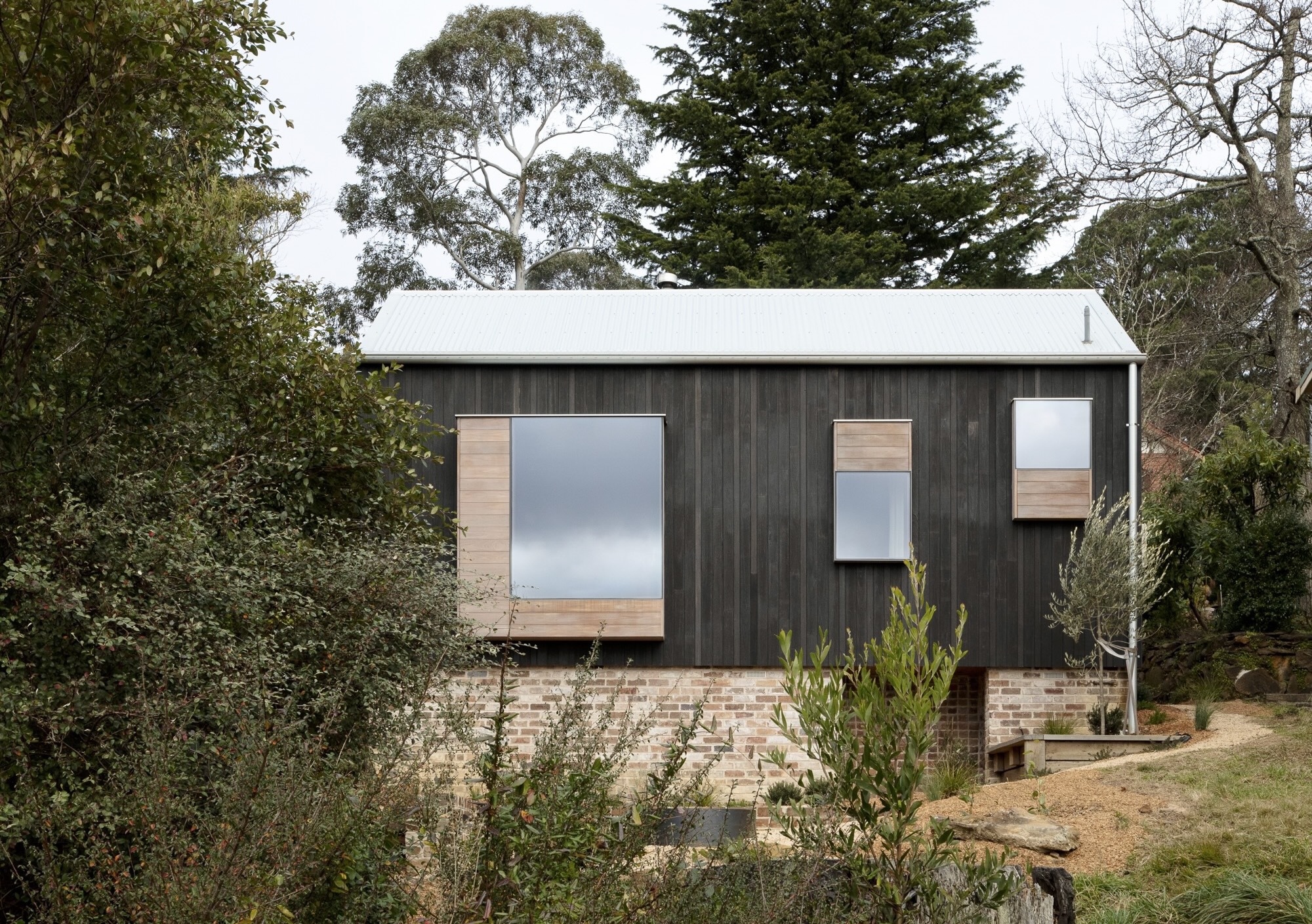 Side view of the dark timber clad volume on brick plinth set into a hillside with eucalyptus trees