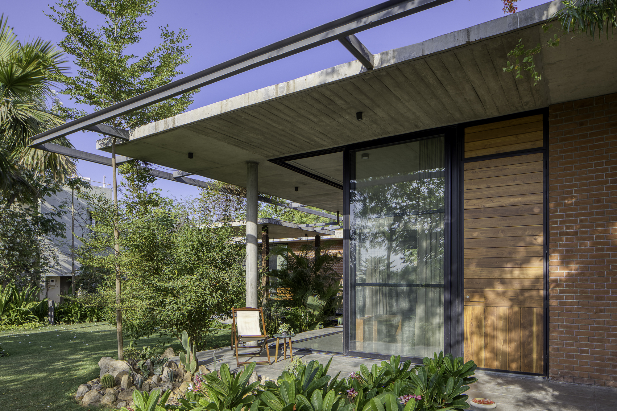 Covered terrace with board-formed concrete ceiling and glass walls framing a planted courtyard with dappled sunlight