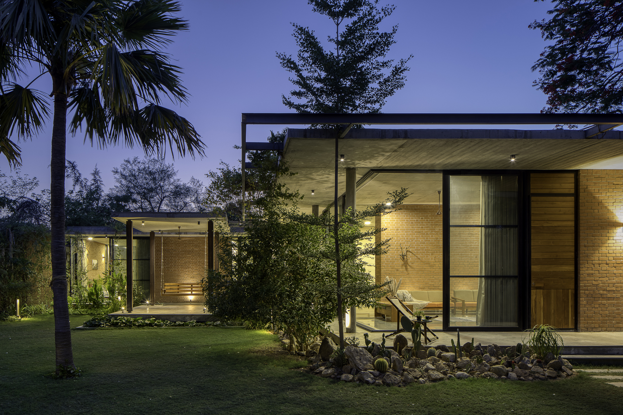 Illuminated glass pavilions with brick and timber facades set in a garden at twilight