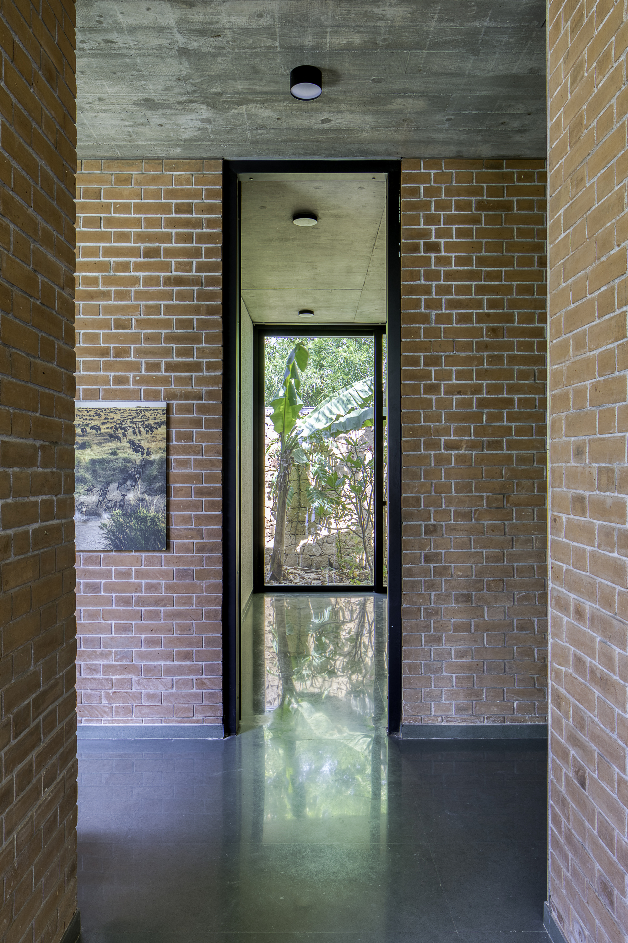 Narrow corridor with buff brick walls flanking a tall vertical window framing vegetation outside