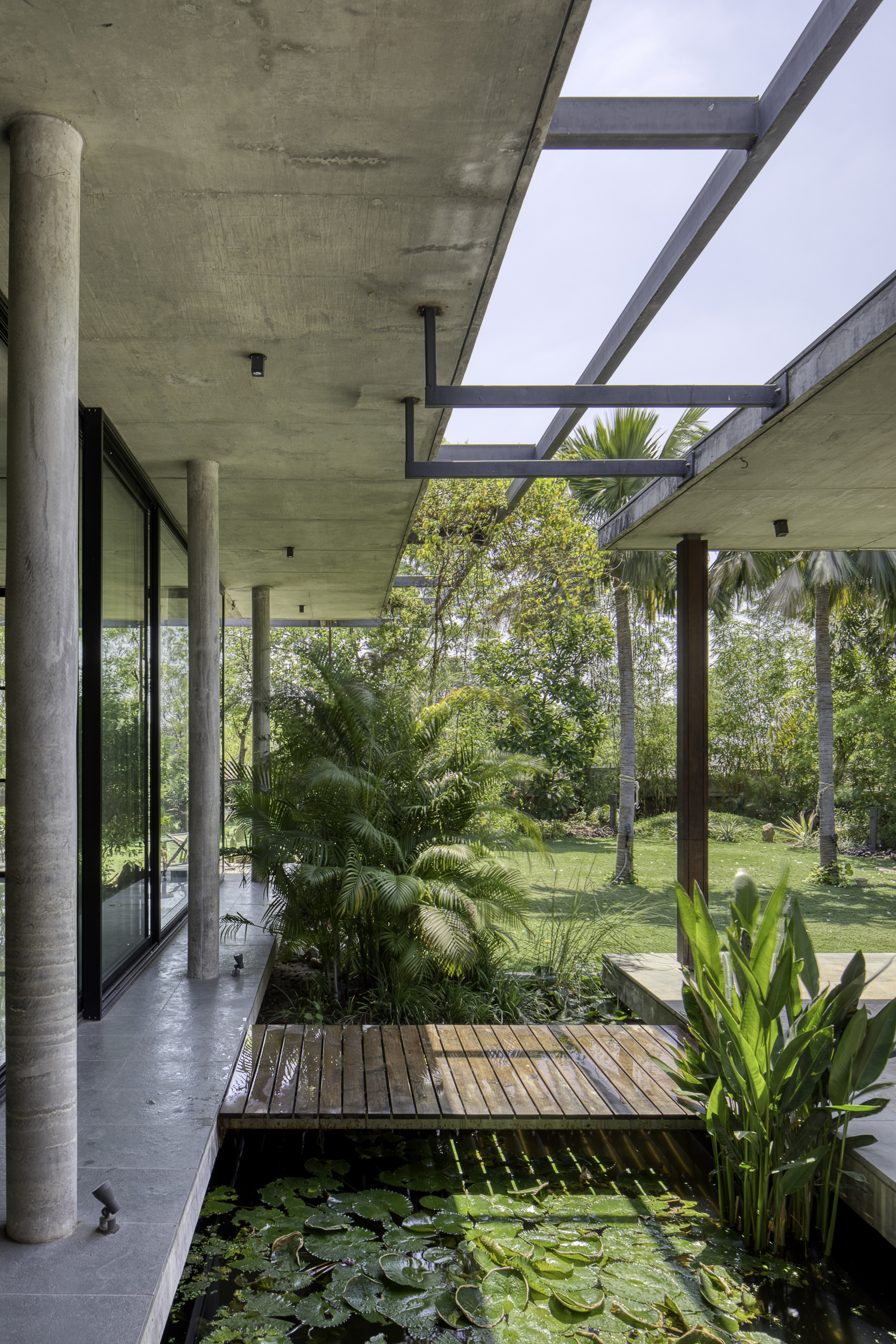 Covered terrace with cylindrical concrete columns and steel pergola overlooking a water garden with palms