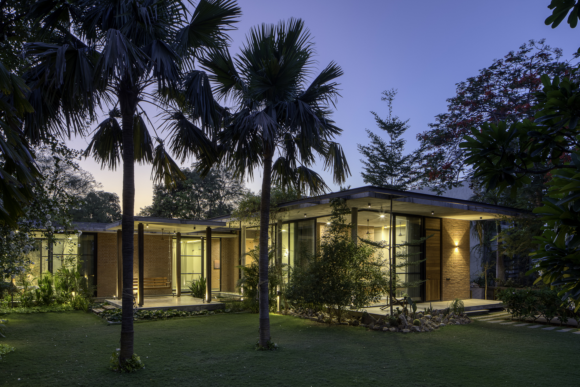 View of the illuminated brick and glass pavilion through palm trees at dusk