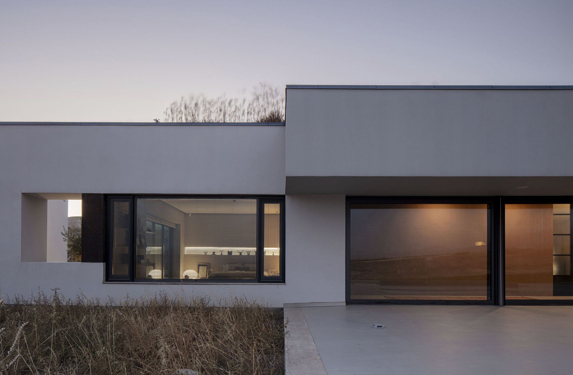 Illuminated interior visible through black-framed glazing at dusk with parched grasses in foreground