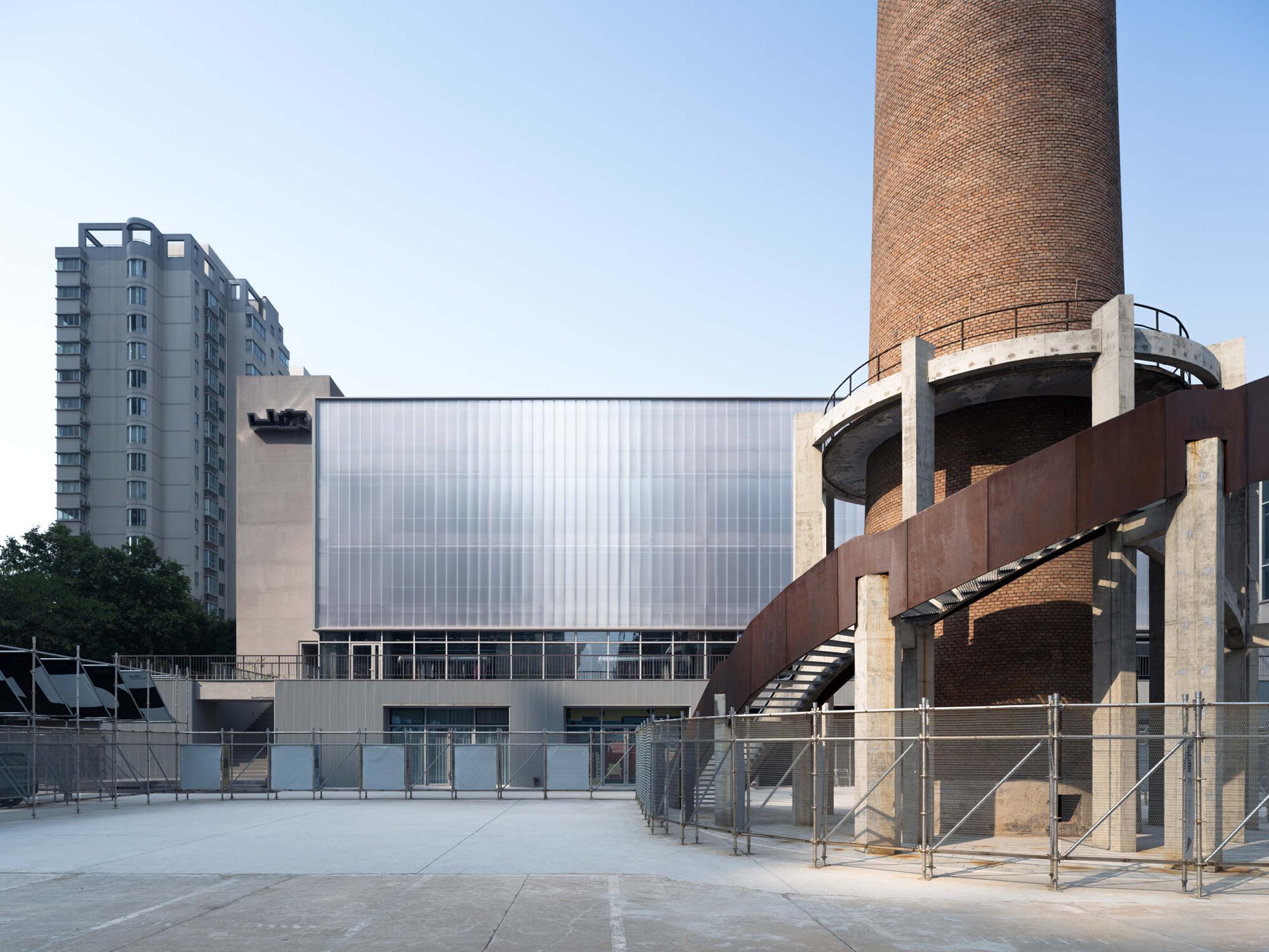 Translucent polycarbonate facade with a weathered brick chimney and elevated steel walkway at dusk