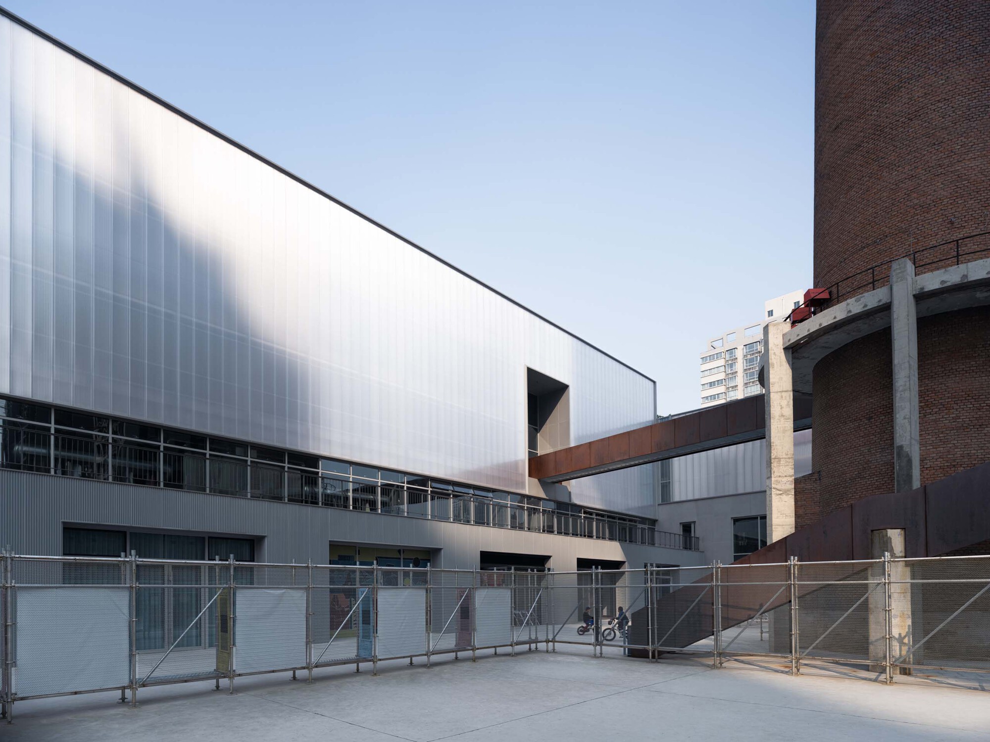 Angular cutout in the translucent facade with weathered steel bridge connecting to the brick chimney structure