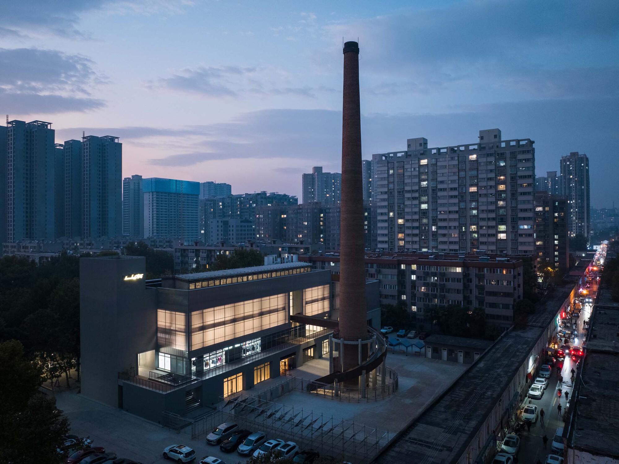 Aerial view of the translucent box volume and tall brick chimney among residential towers at dusk