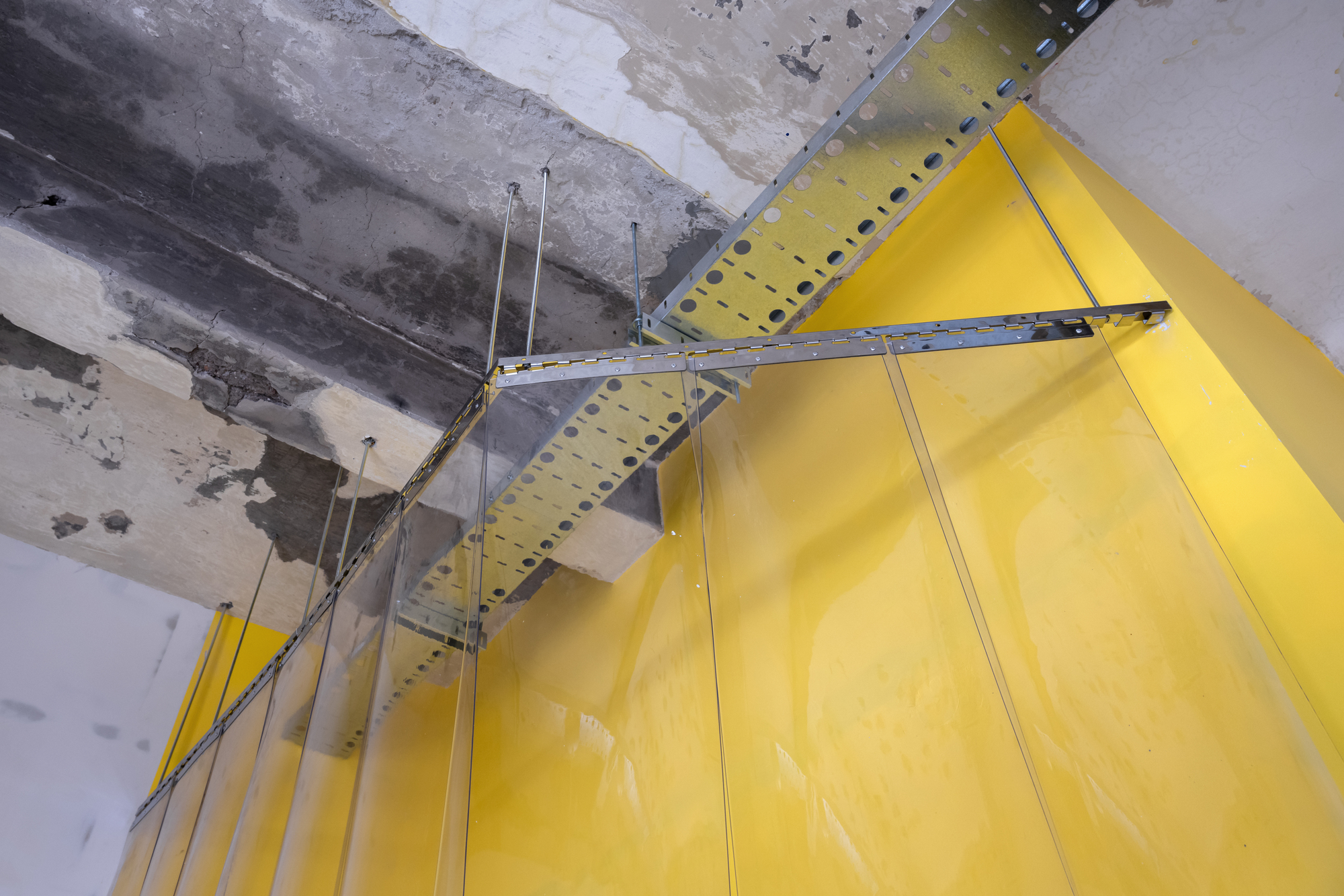 Yellow curved partition wall suspended from exposed concrete ceiling with metal brackets and rods
