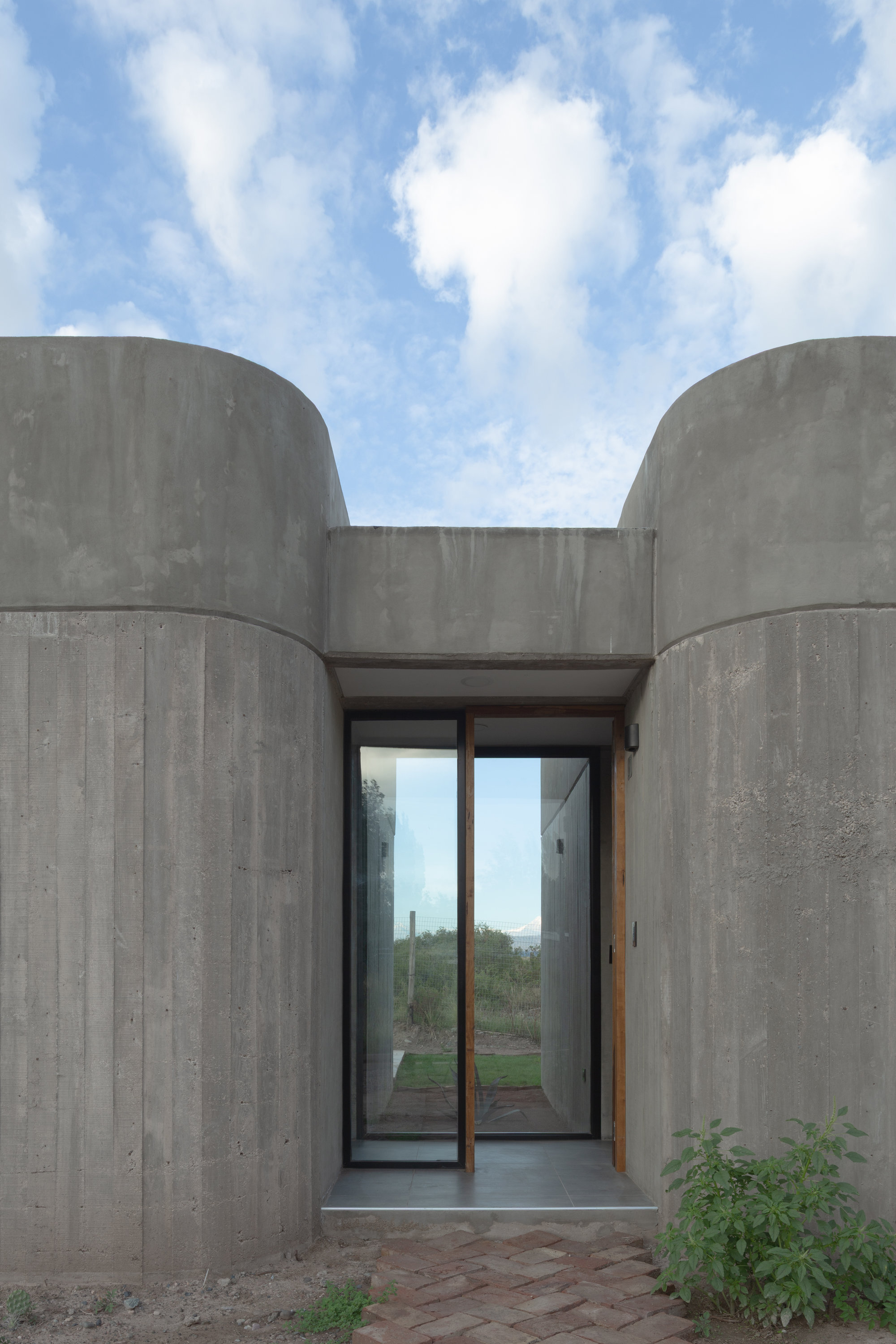 Glass entrance doors set between two cylindrical board-formed concrete volumes under a blue sky
