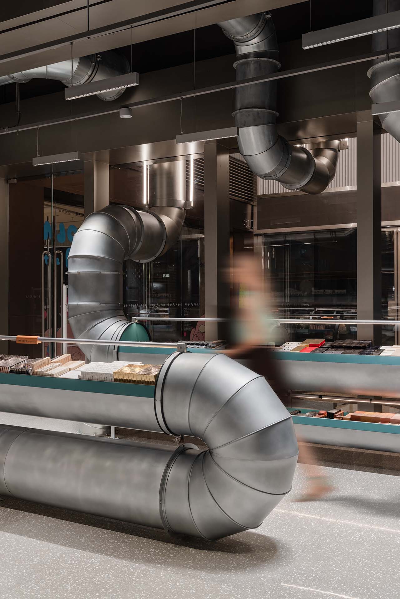 Curving metal ventilation duct crossing the retail floor as a blurred figure passes behind