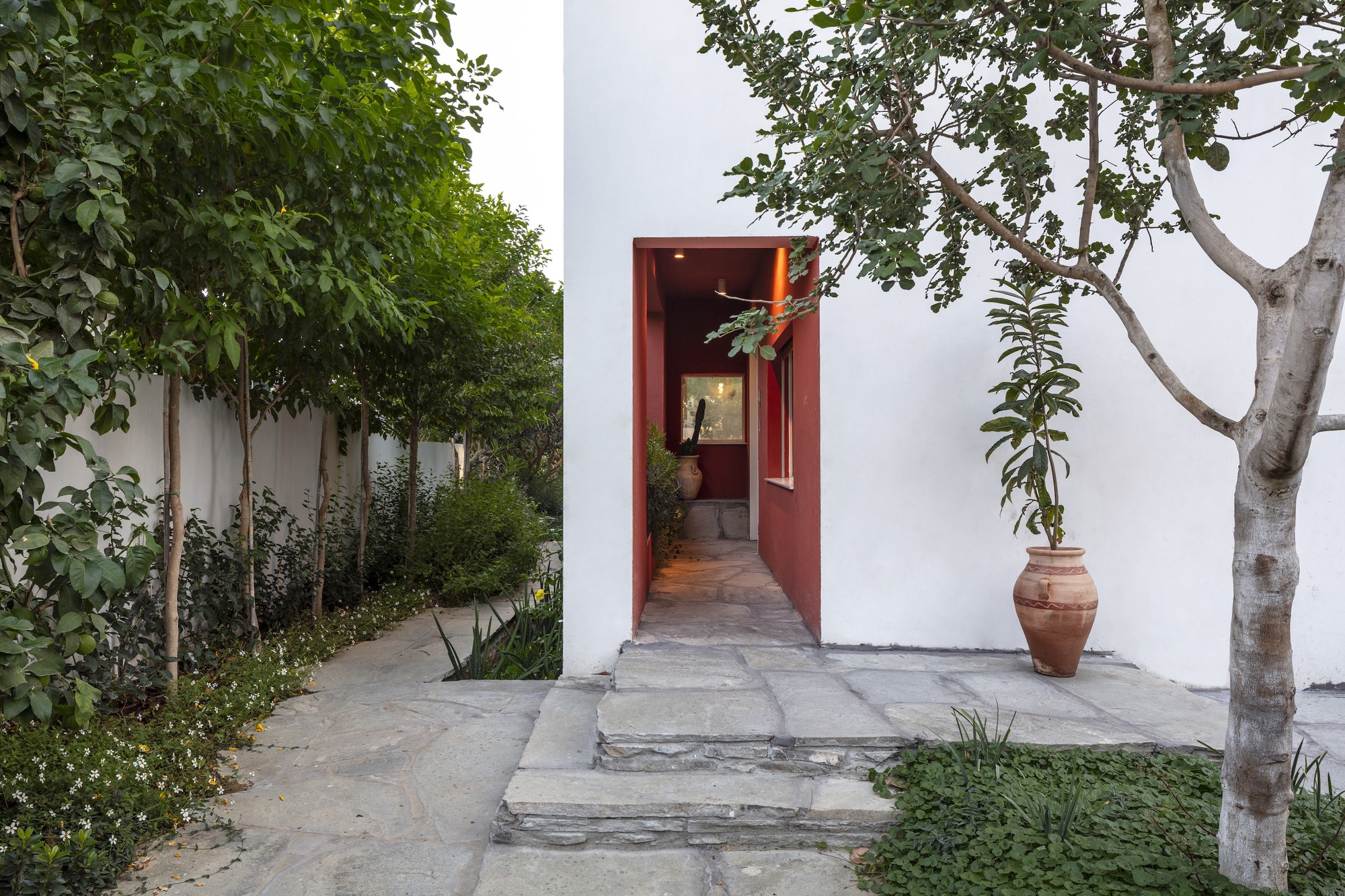 White stucco courtyard with red-framed doorway flanked by planted beds and terracotta pot