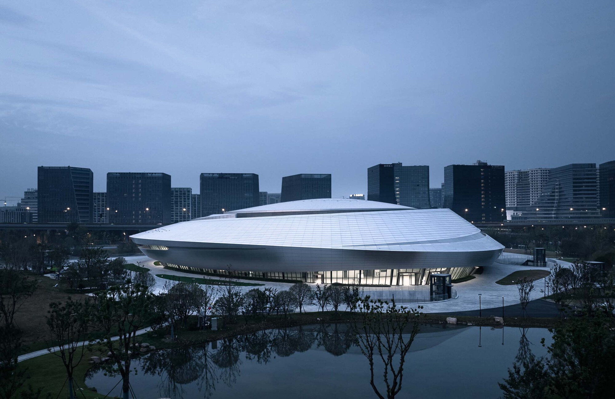 Ribbed white roof cantilevering over a glazed entry plaza at dusk with trees and water in the foreground