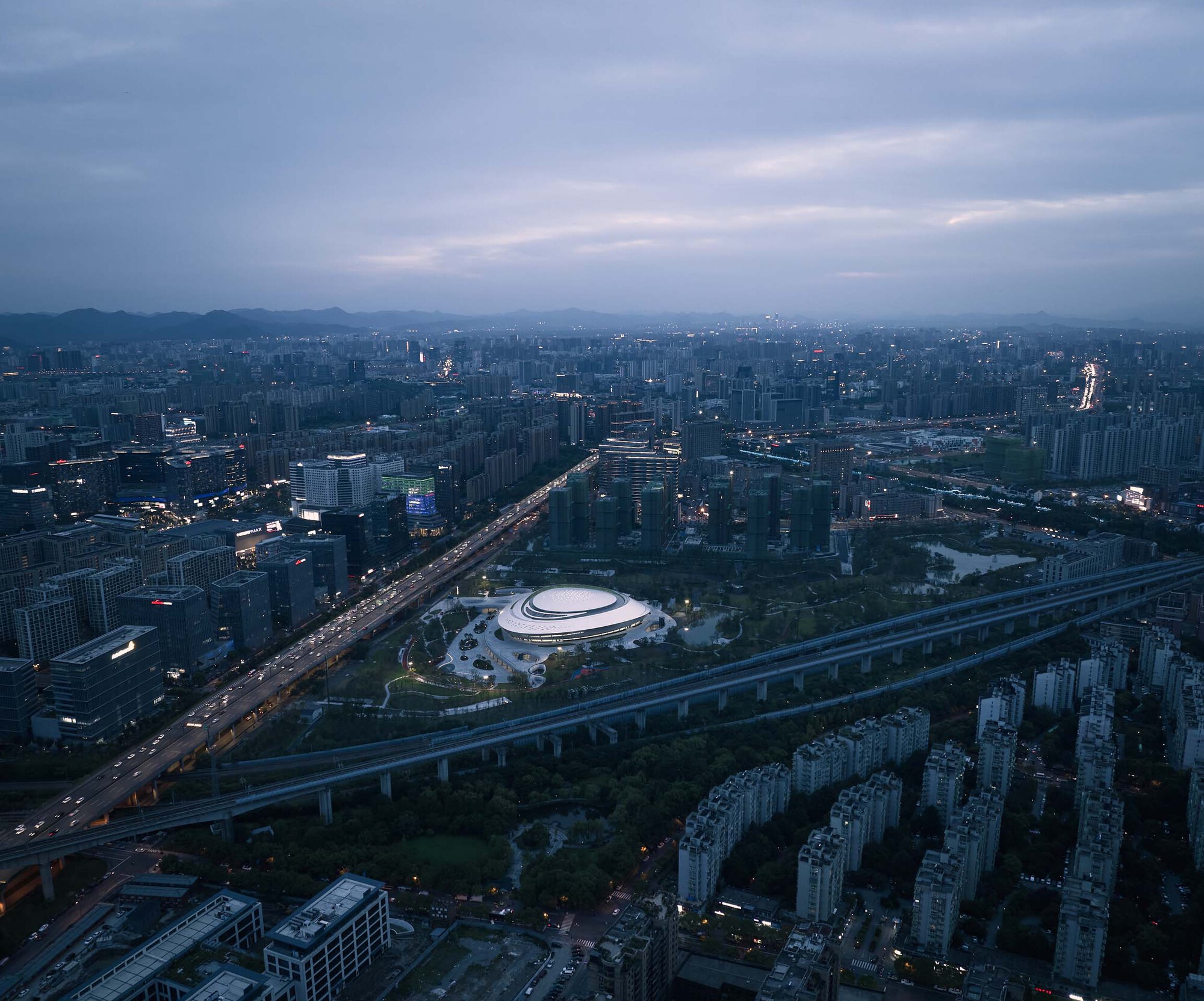 High-altitude view of the building nestled between elevated roads and residential towers at dusk