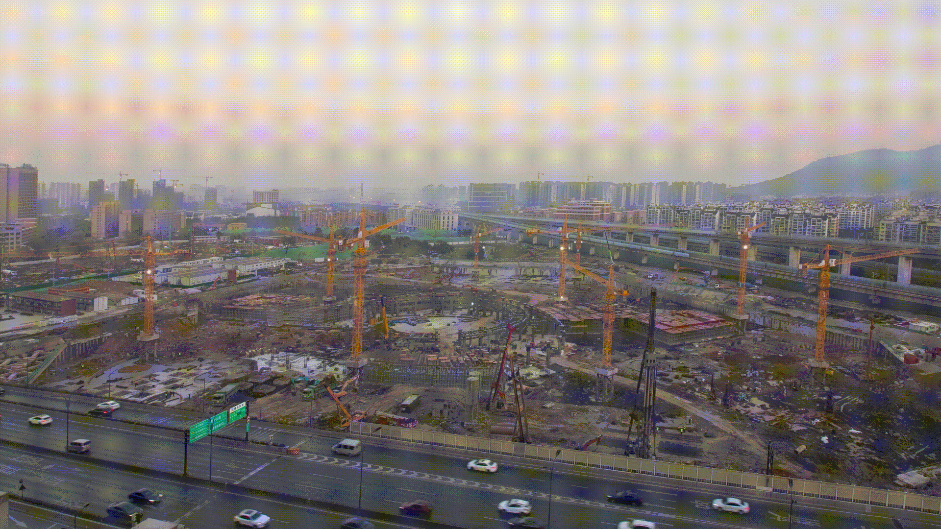 Aerial view of the construction site with yellow cranes and surrounding urban infrastructure at dusk