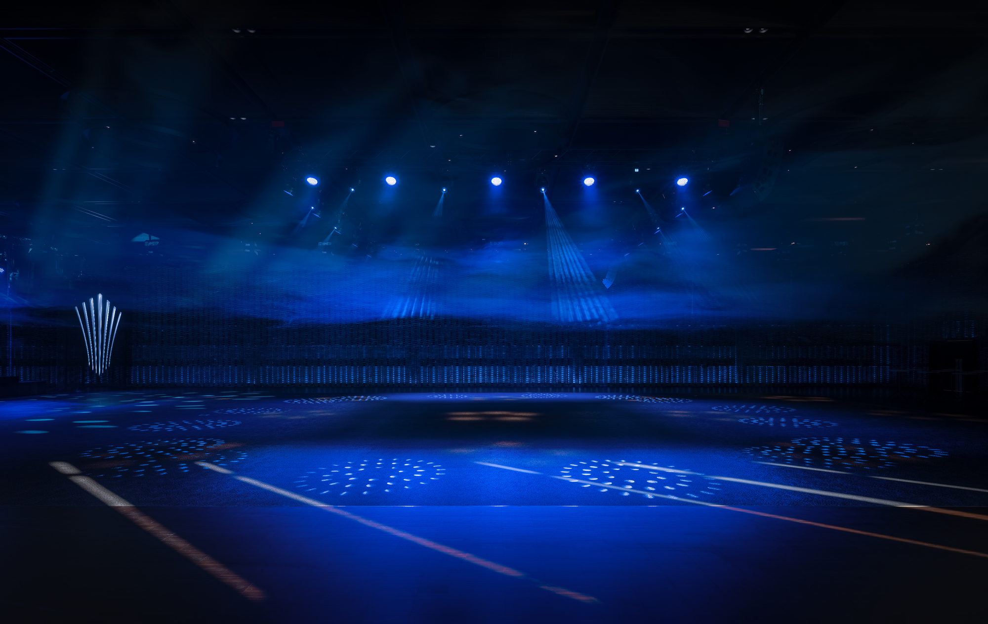 Interior arena space with blue stage lighting and perforated ceiling visible through atmospheric haze