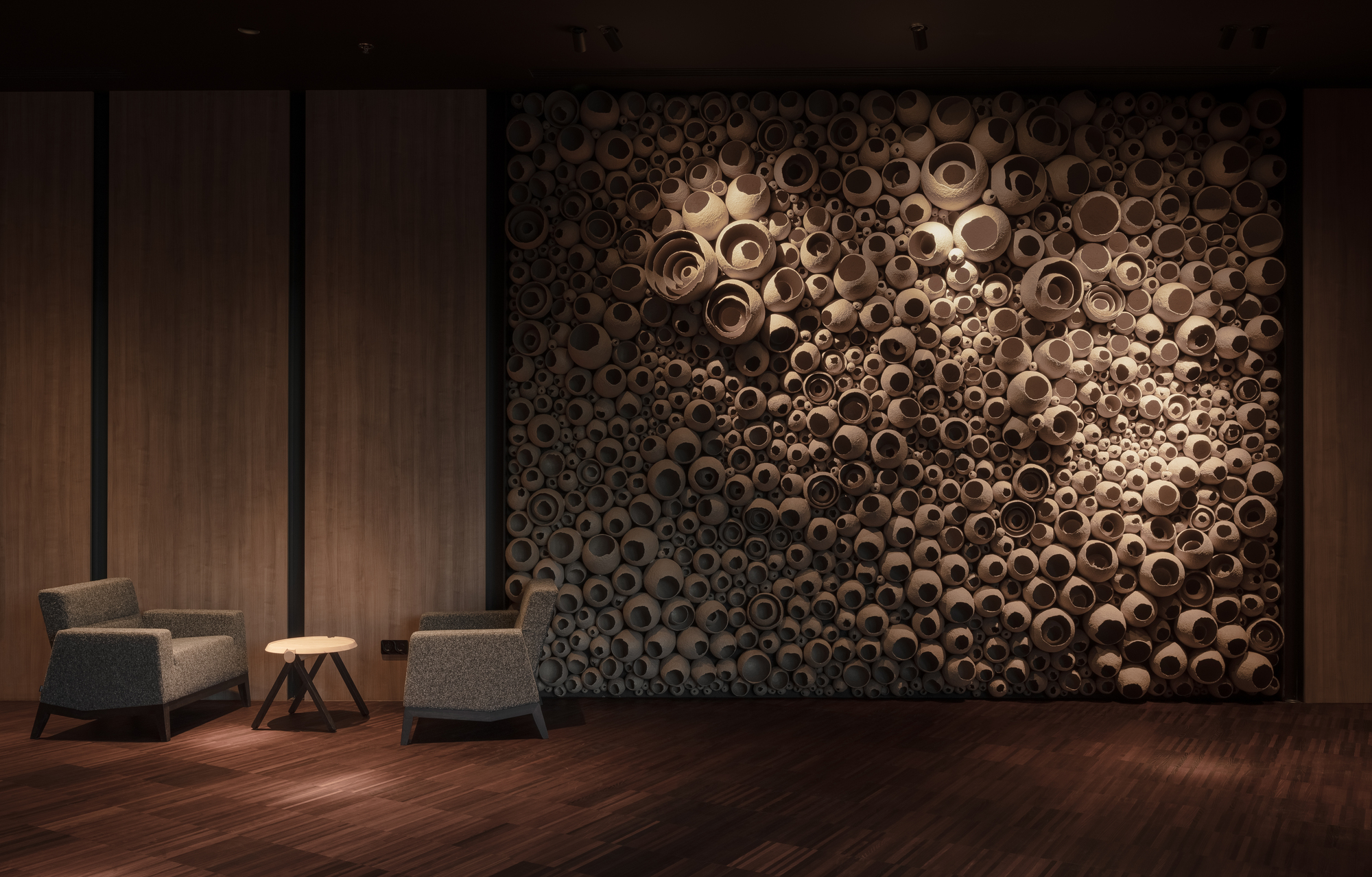 Sculptural wall installation of clustered cylindrical timber tubes backlit against wood paneling with two lounge chairs
