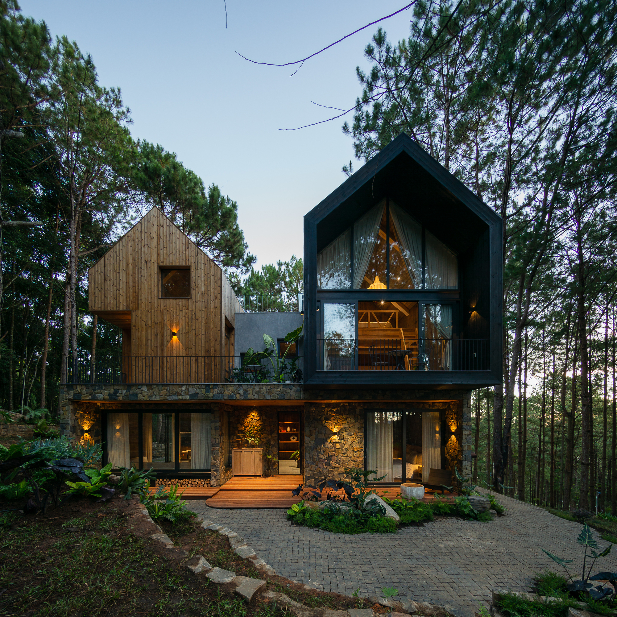 Front facade showing timber and dark metal gabled volumes above a stone base at dusk
