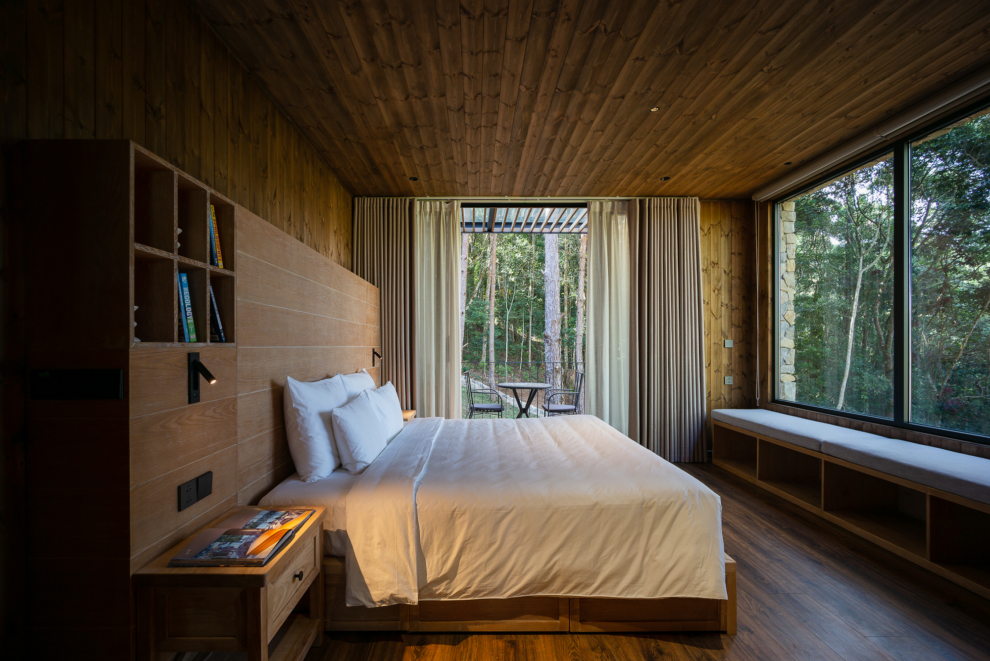 Bedroom opening to a glazed balcony with forest views through floor-to-ceiling windows and curtains