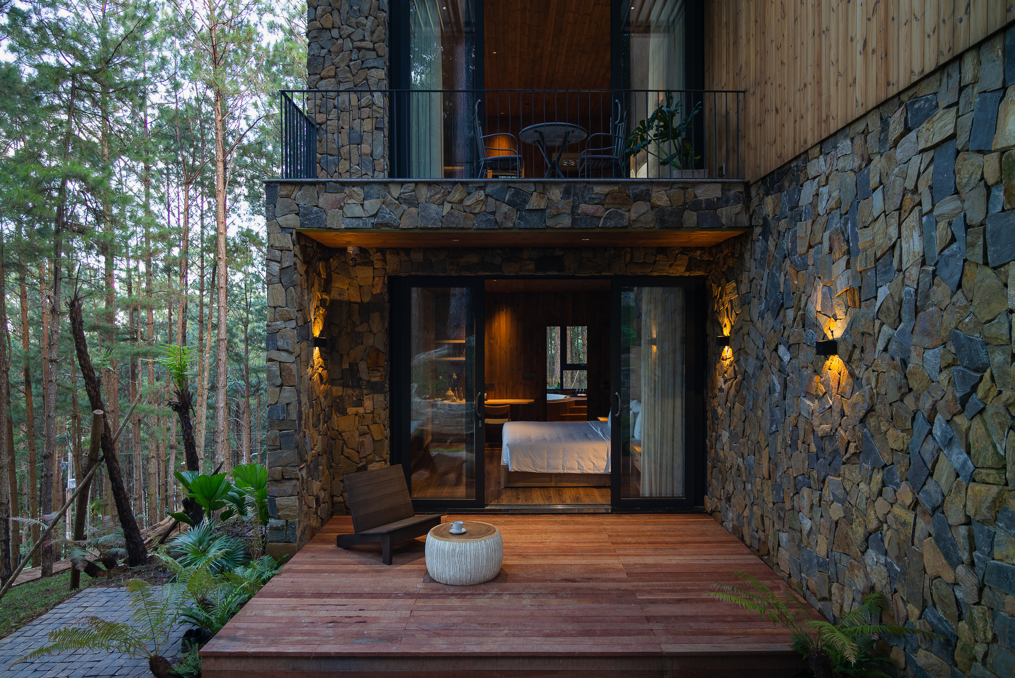 Timber deck terrace leading to glazed bedroom entry framed by stacked stone walls and forest