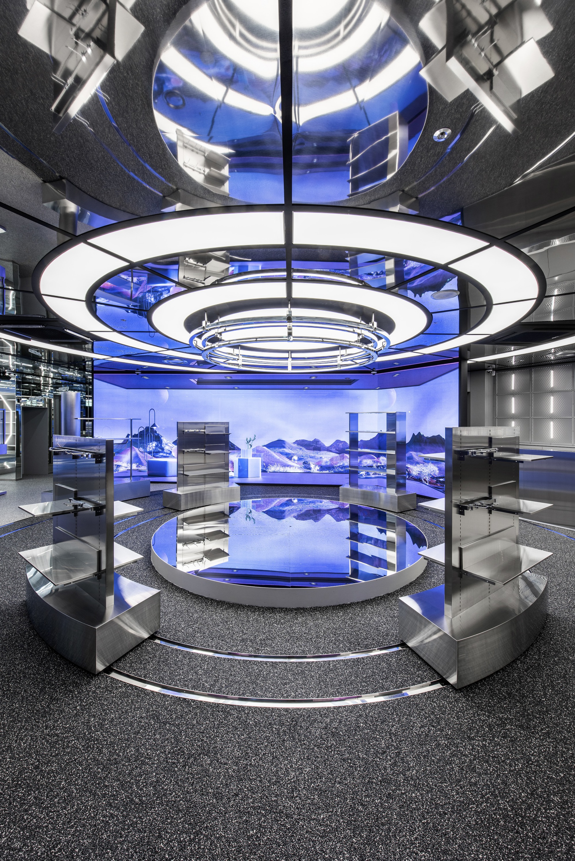 Tiered display platforms beneath concentric ceiling rings reflected in polished floor with blue projection screens beyond
