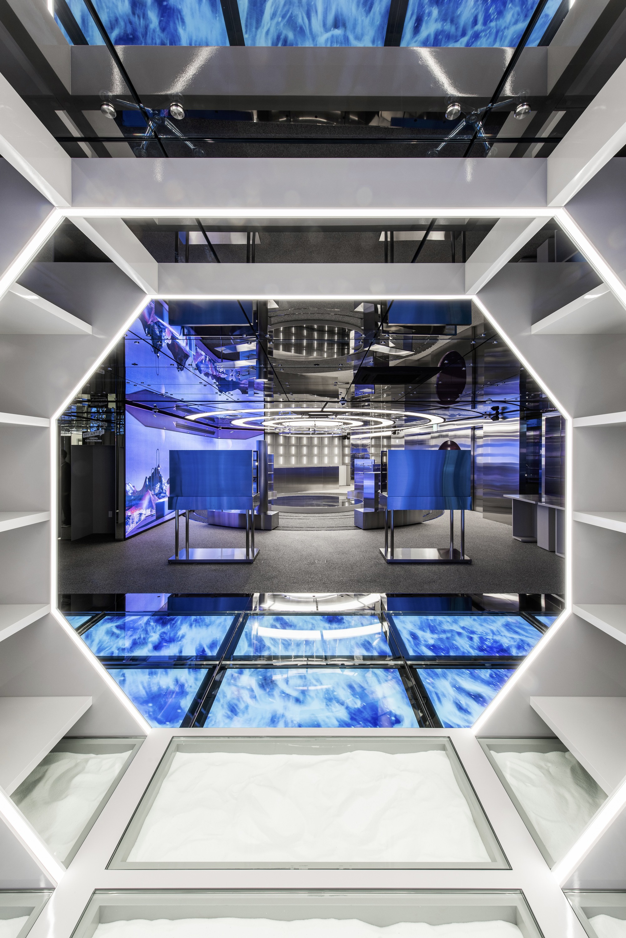 Looking down through octagonal void with glass floor panels and blue digital screens