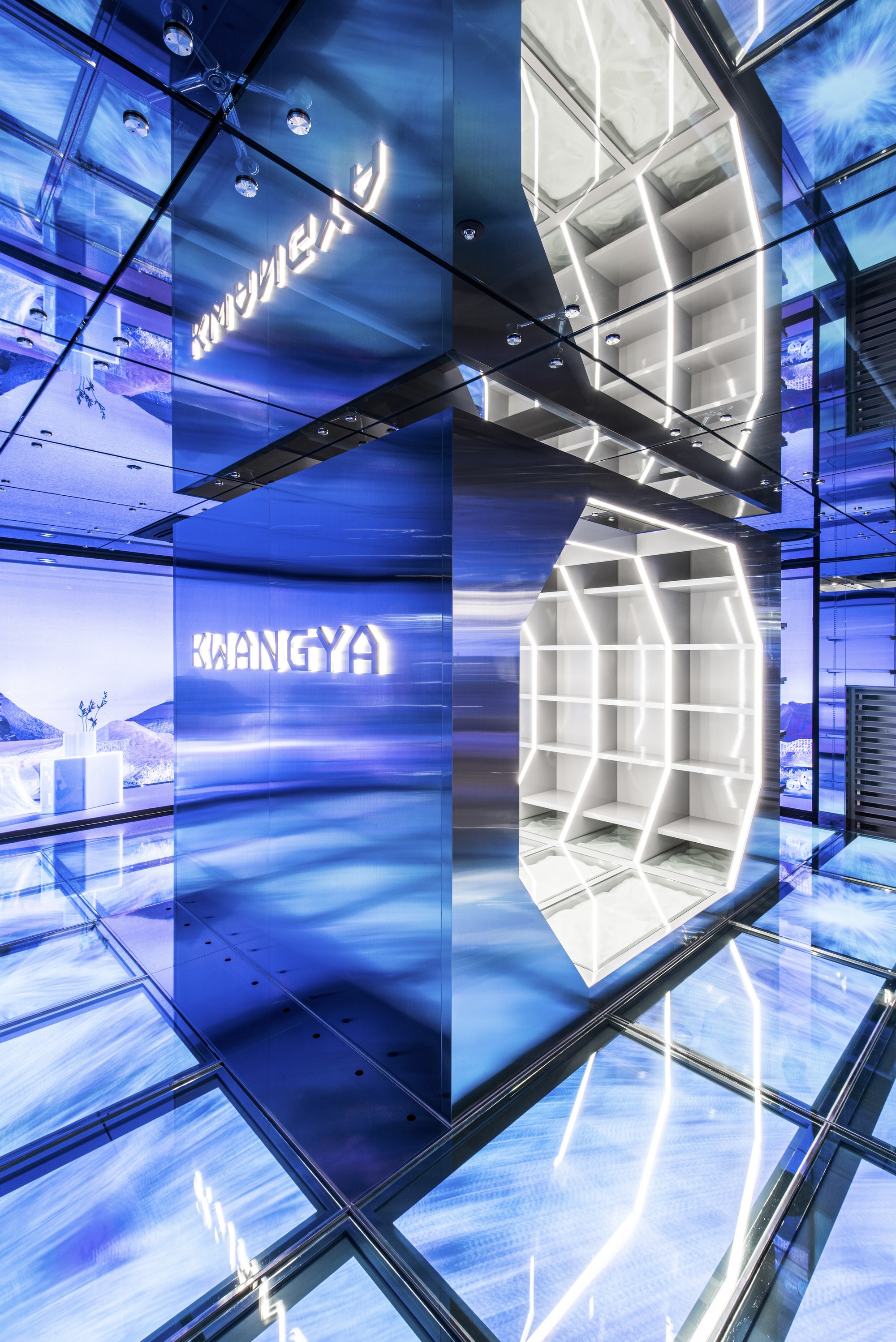 Multi-story retail atrium with illuminated blue glass walls and suspended white display cube under glass floor