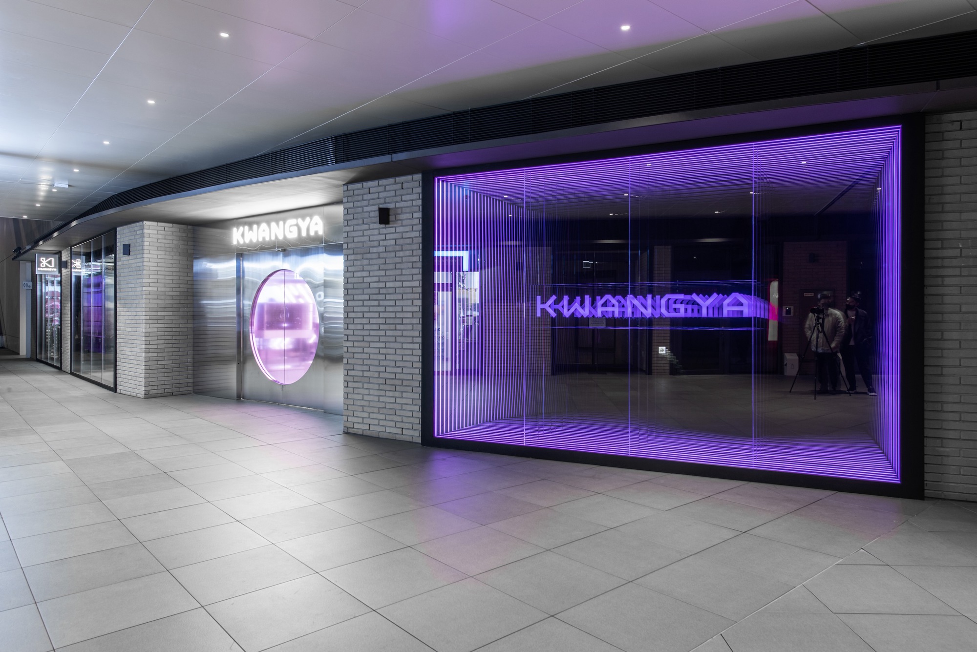 Retail storefront with purple LED-lit glass facade and grey brick walls along a mall corridor