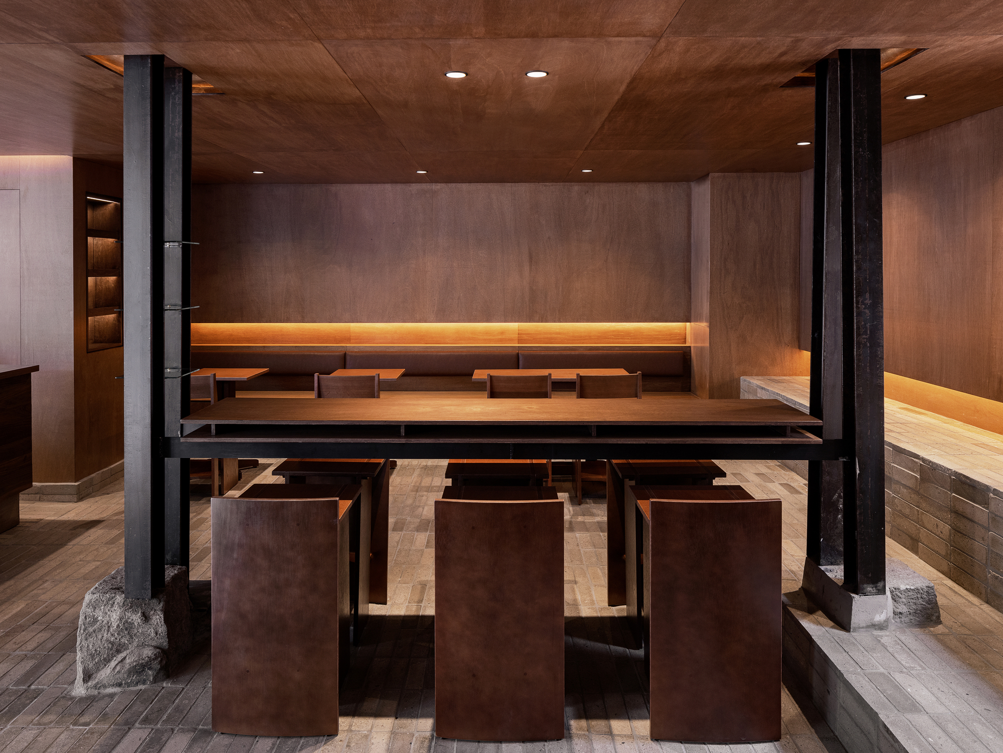 Dining area with long timber tables, metal cube stools, and black steel columns beneath a wood ceiling