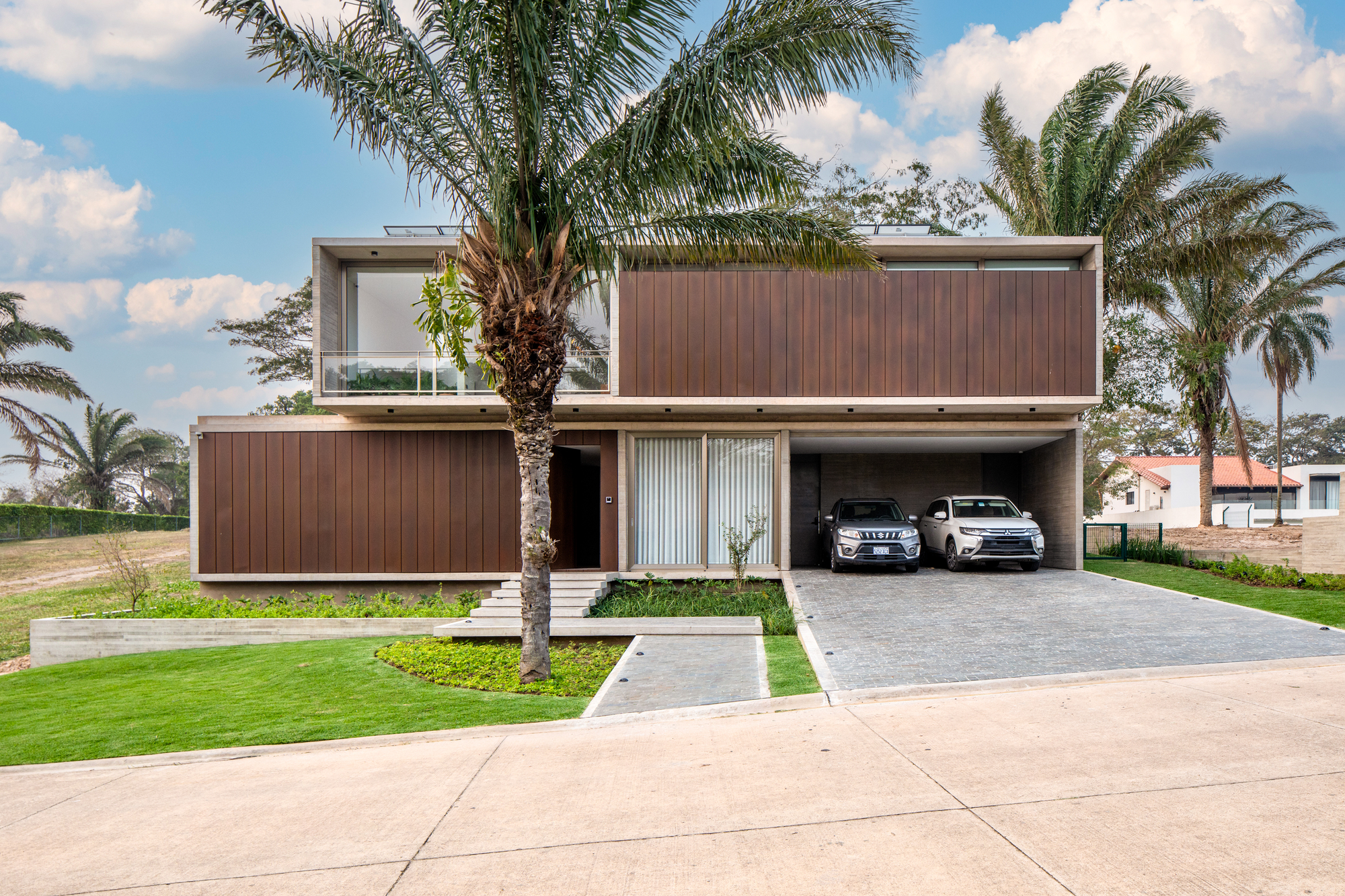 Street facade with vertical timber cladding, open carport, and palm trees under a cloudy sky