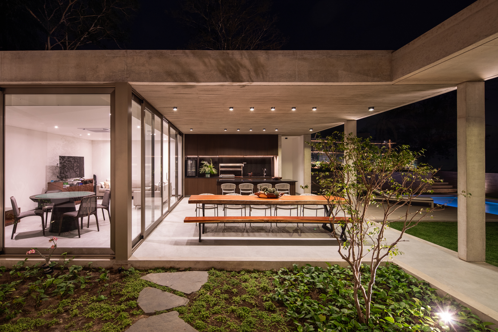 Covered dining terrace with concrete frame opening to planted courtyard and pool at night