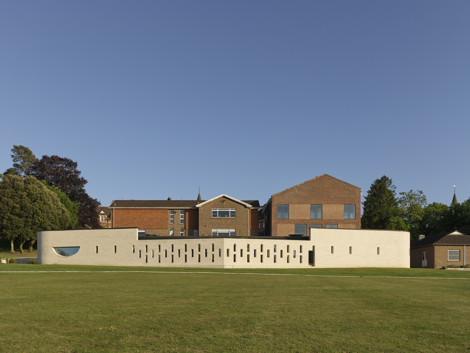 Pale concrete wall with vertical slit windows facing a green cricket pitch under clear blue sky