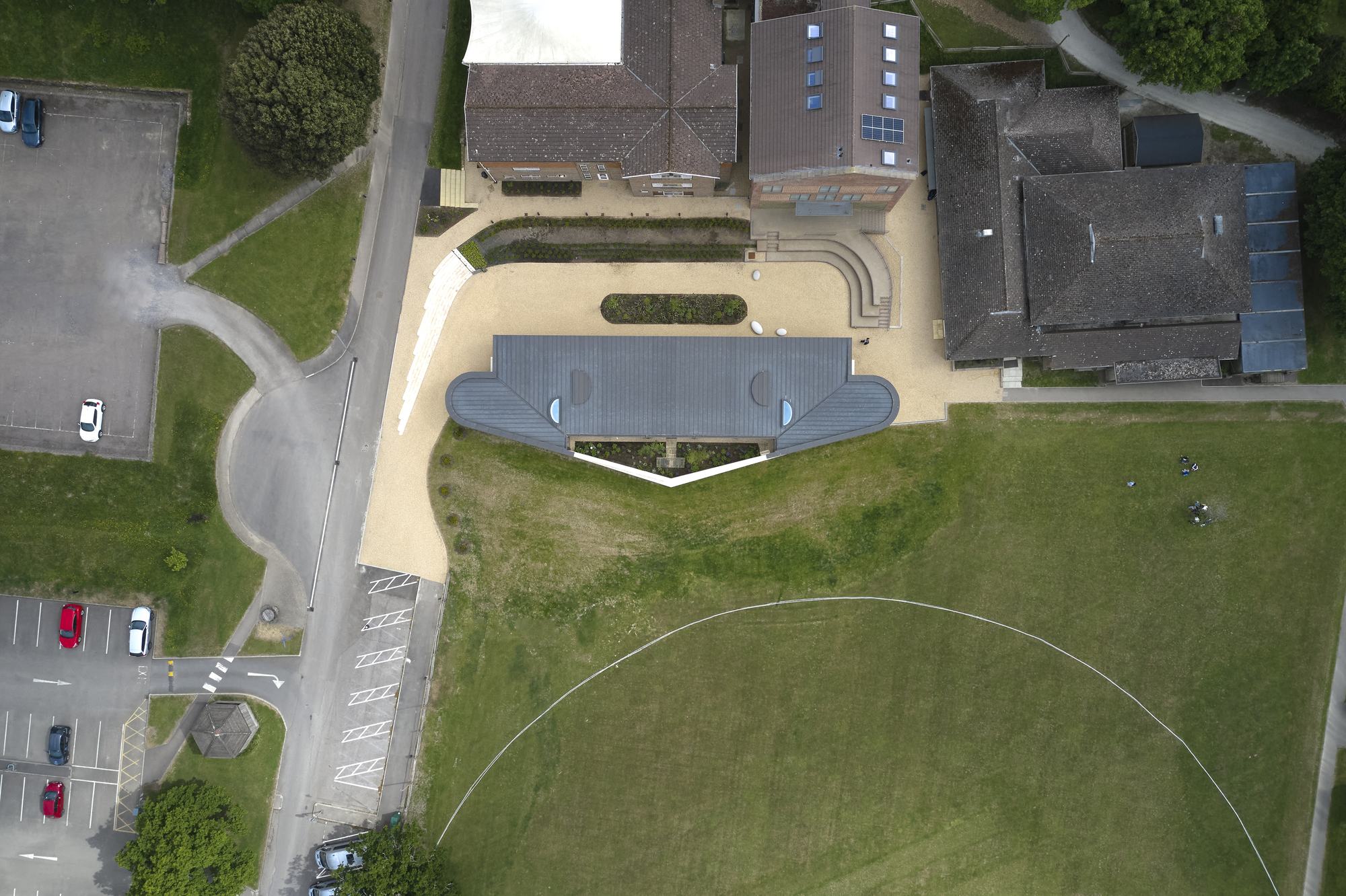 Aerial view of the curved pavilion with dark metal roof surrounded by gravel courtyard and grass